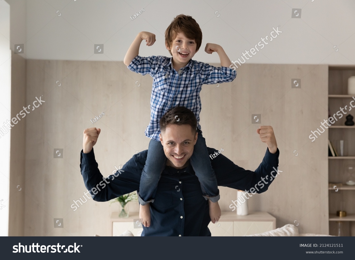 Cheerful little boy riding on happy dads neck and shoulders, making powerful hands. Strong daddy keeping balance with kid on top, flexing arm muscles, smiling at camera. Father and son portrait #2124121511