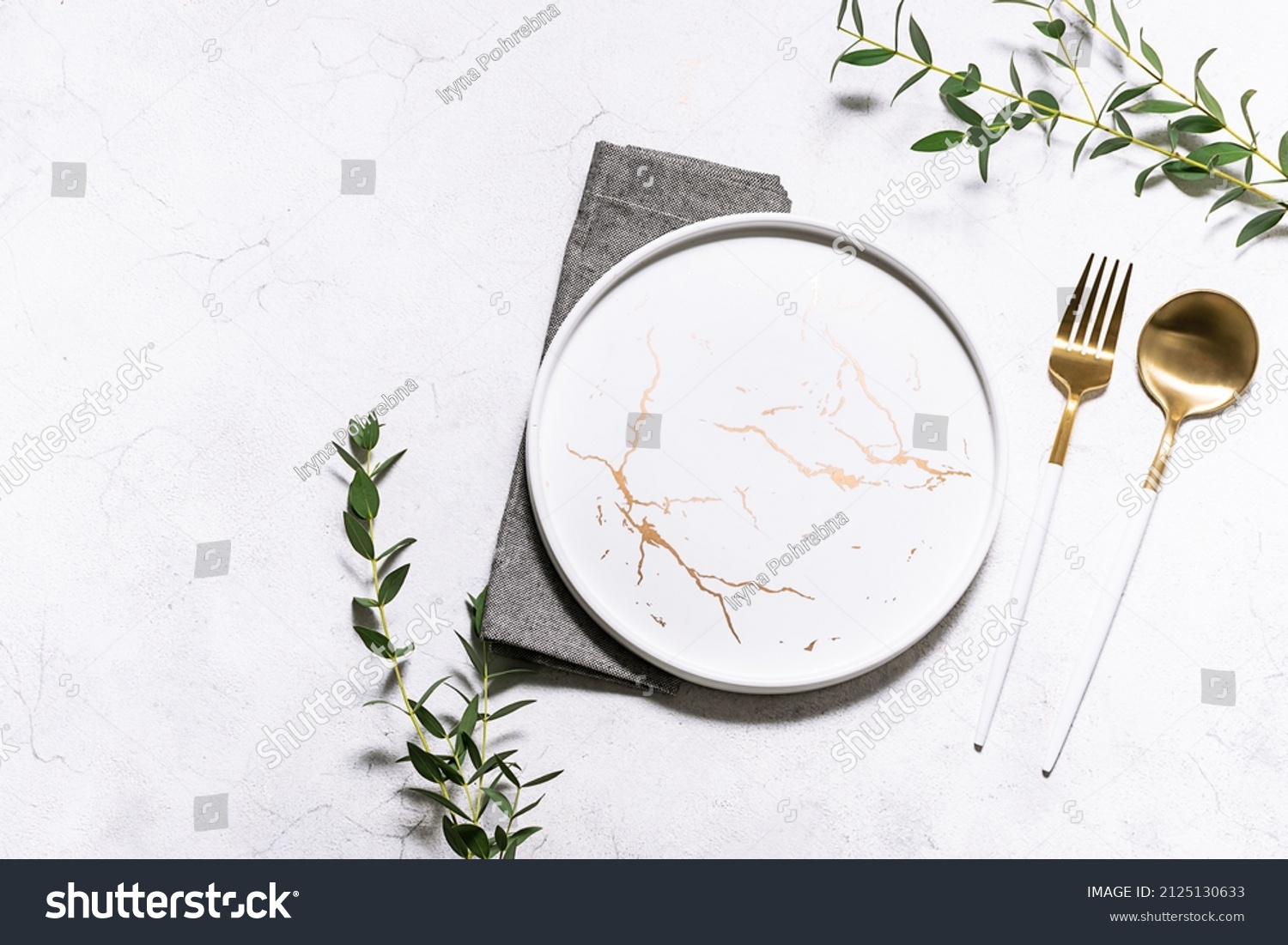 Festive table setting with white marble plate, golden cutlery set and fresh eucalyptus leaves on white rustic table top view. Copy space for your design. #2125130633