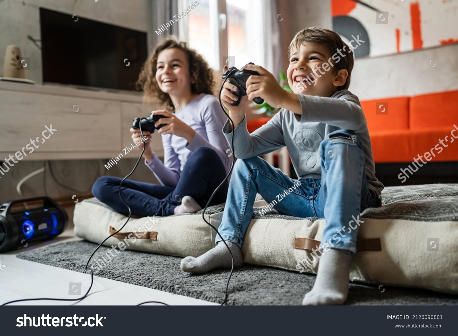 Two children small caucasian brother and sister happy children siblings boy and girl playing video game console using joystick or controller while sitting at home real people family leisure concept #2126090801
