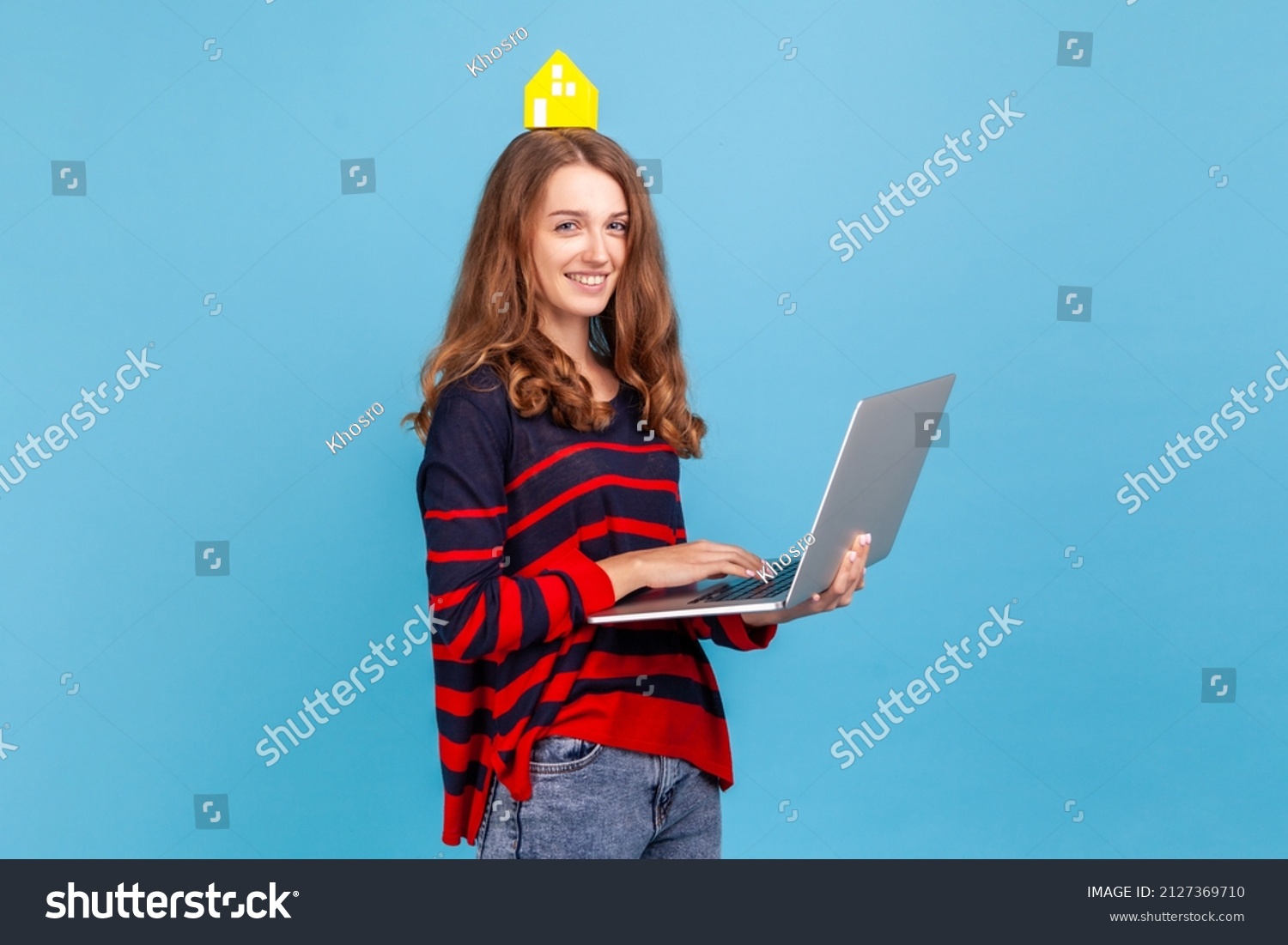 Attractive happy woman wearing striped casual style sweater, holding laptop in hands, looking at camera, holding paper house on head, online renting. Indoor studio shot isolated on blue background. #2127369710