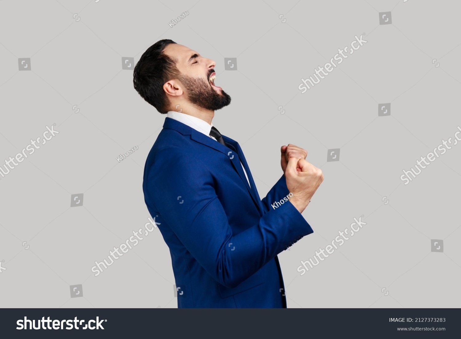 Side view of bearded man showing yes gesture and screaming celebrating his victory, success, dreams comes true, euphoria, wearing official style suit. Indoor studio shot isolated on gray background. #2127373283