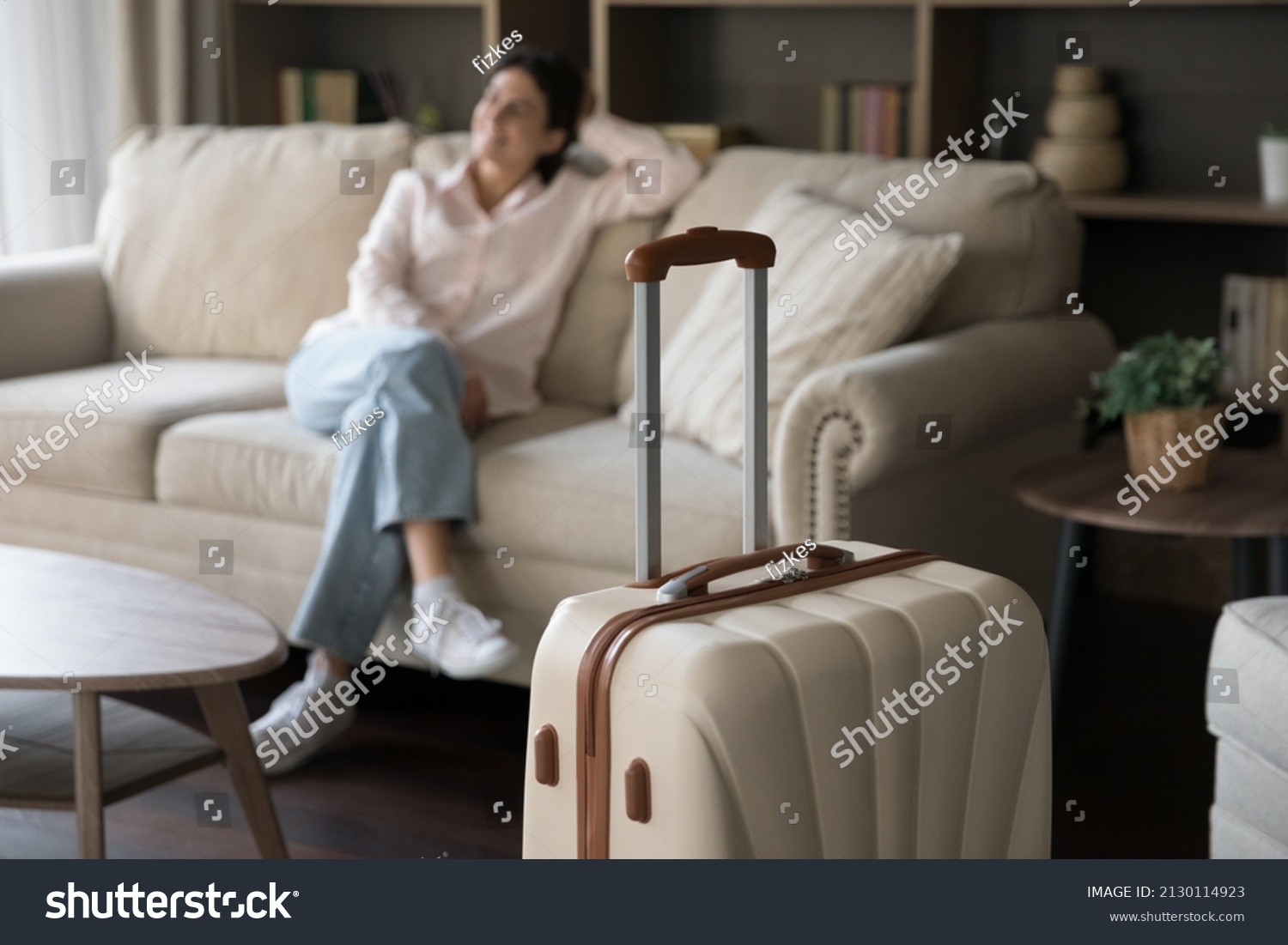 Focus on beige suitcase with blurred carefree young woman sitting on sofa on background. Joyful refreshed millennial Hispanic lady satisfied with vacation travel, relaxing alone in modern hotel room. #2130114923