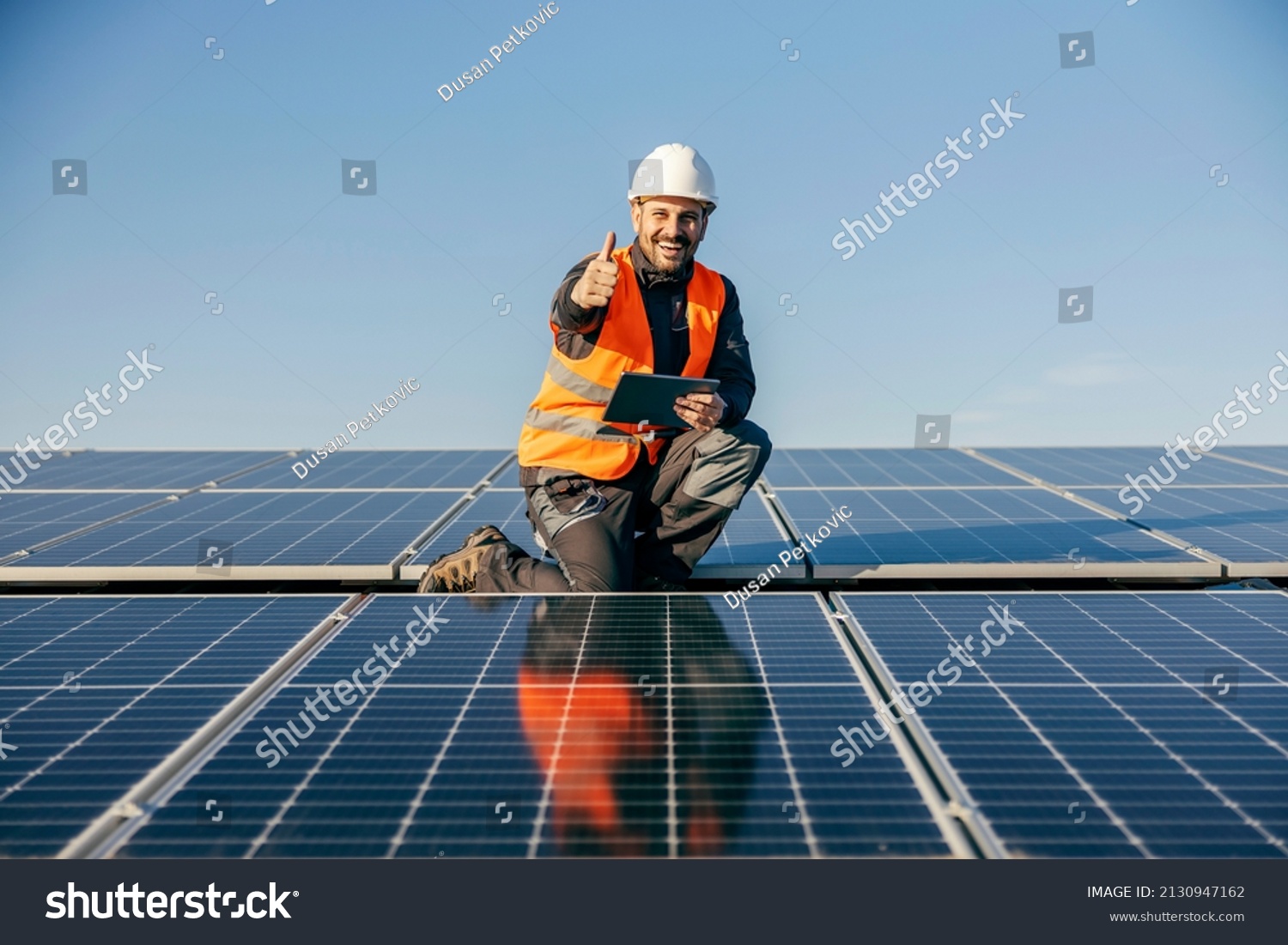 A worker on rooftop kneeling next to solar panels with tablet in hands and giving thumb up for sustainable life. #2130947162