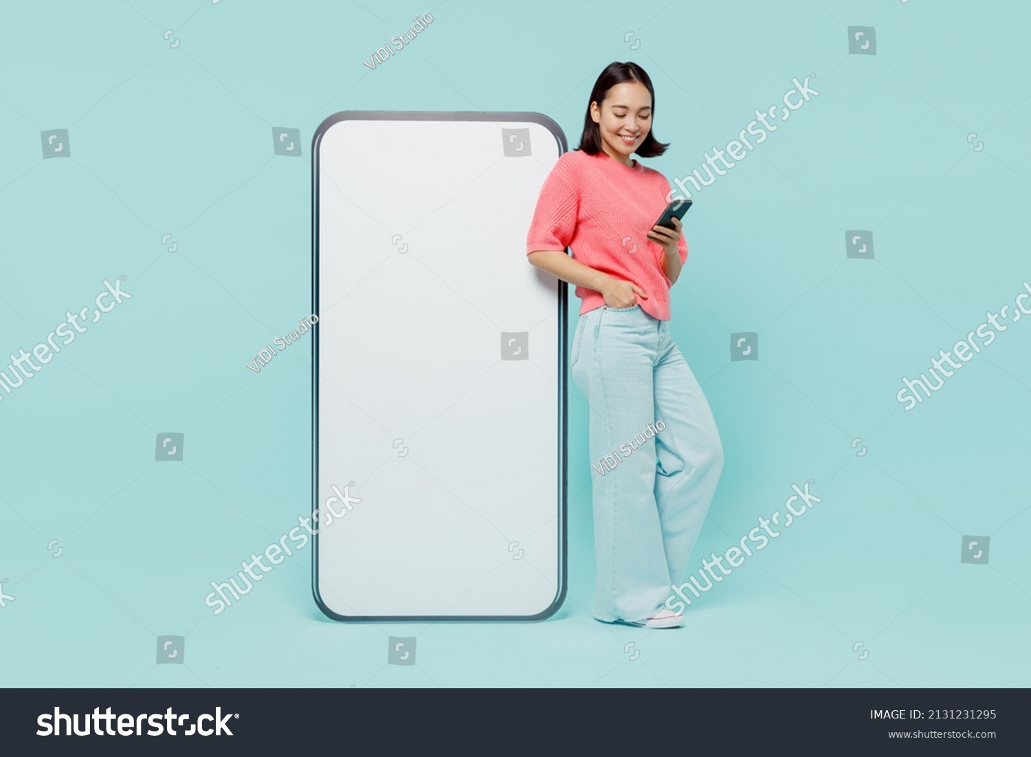 Full body young smiling happy woman of Asian ethnicity 20s in pink sweater stand near big mobile cell phone with blank screen workspace area chatting isolated on pastel plain light blue background. #2131231295