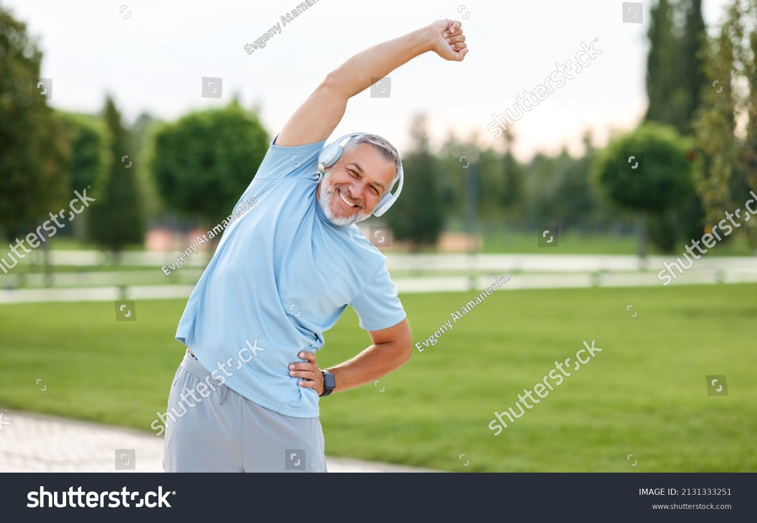 Staying active after retirement. Happy joyful mature retired sportsman wearing headphones and sportswear doing side stretching exercises with arm over his head, exercising outside in city park #2131333251