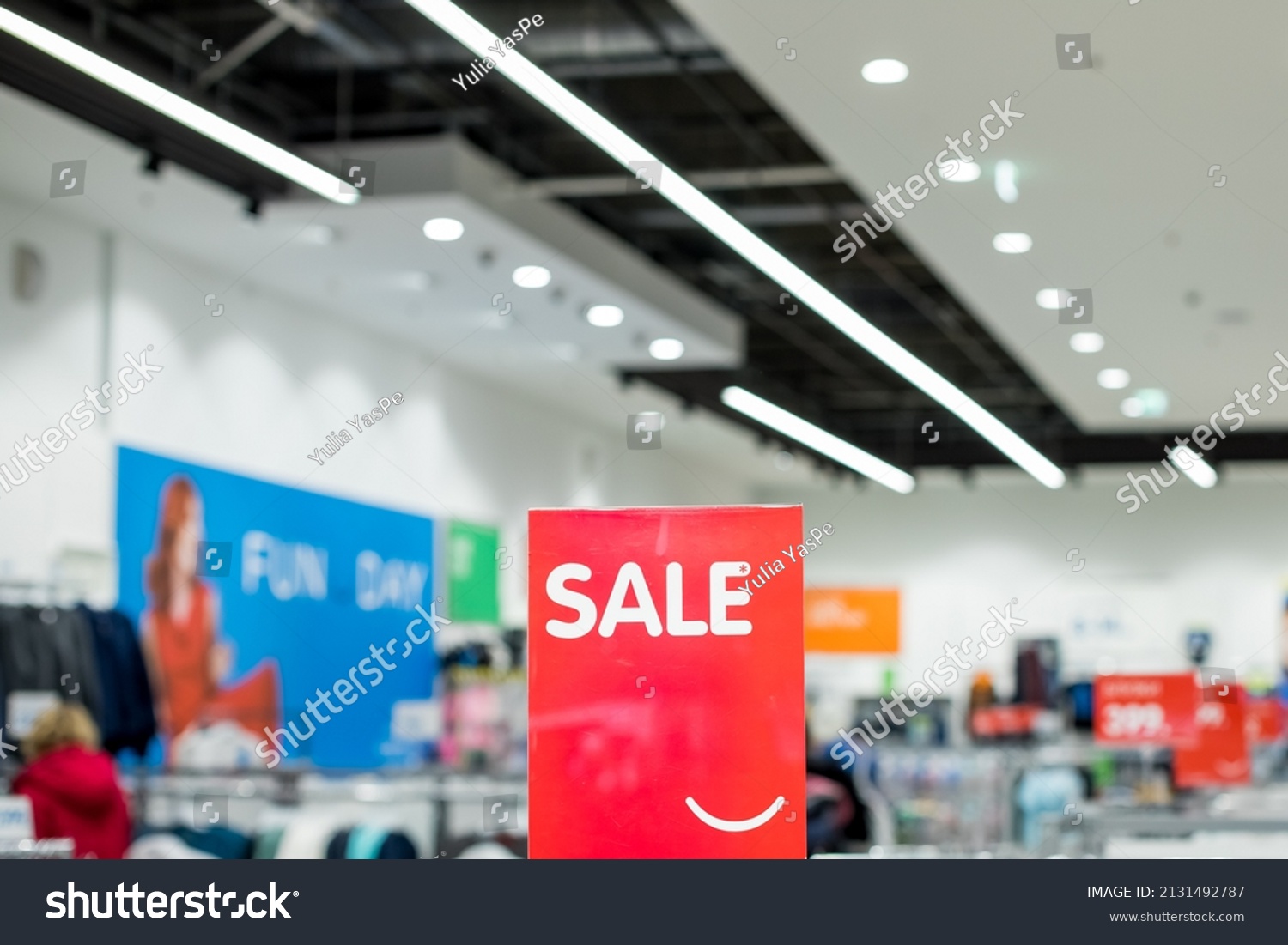 A sign with the inscription SALE in white on a red background in the trading floor of casual clothing store. Fashion concept, discount season, black friday, offline shopping, gimmicks, holiday sales. #2131492787