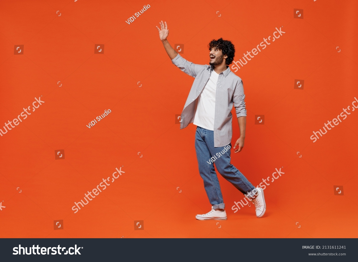 Full size body length side profile view cheerful young bearded Indian man 20s years old wears blue shirt meet greet waving hand as notices someone isolated on plain orange background studio portrait #2131611241
