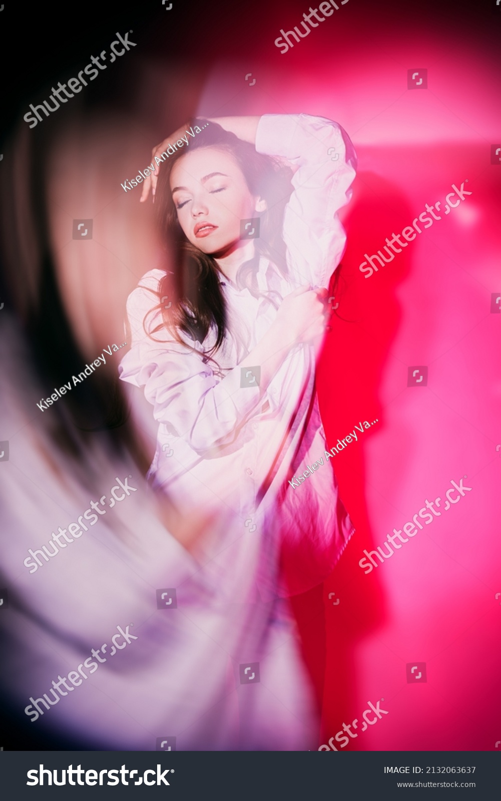 A young beautiful girl is standing with closed eyes in euphoria. The face is illuminated by a beam of light. Thoughtfulness, insight. Studio portrait in crimson lighting. #2132063637