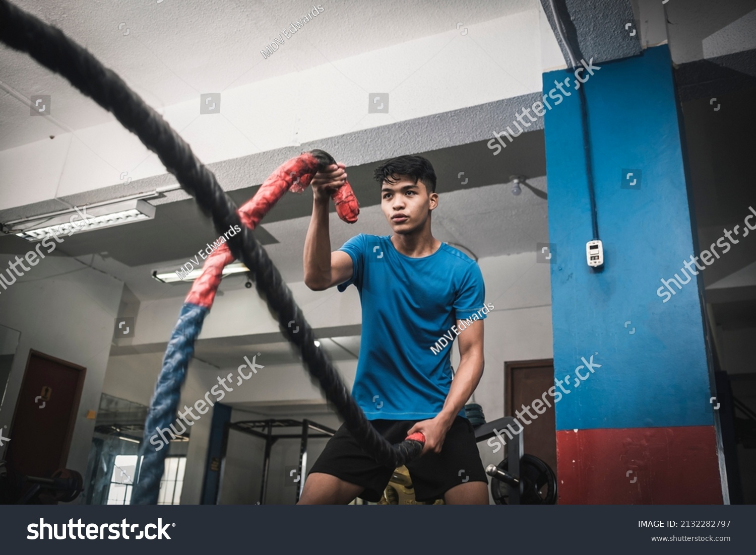A fit young asian man working out vigorously with battle ropes. Alternating single arm waves. Whole body workout, conditioning and cardio at the gym. #2132282797