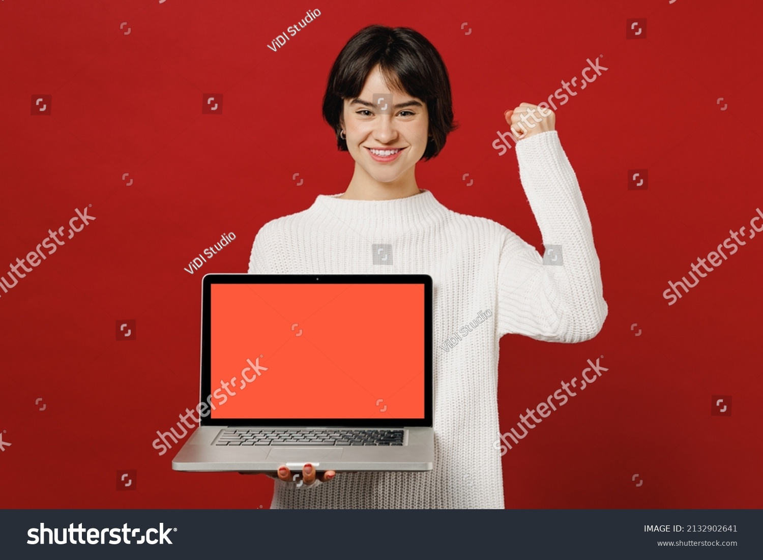 Young happy fun excited woman 20s wearing white knitted sweater holding use work on laptop pc computer with blank screen workspace area do winner gesture isolated on plain red color background studio. #2132902641