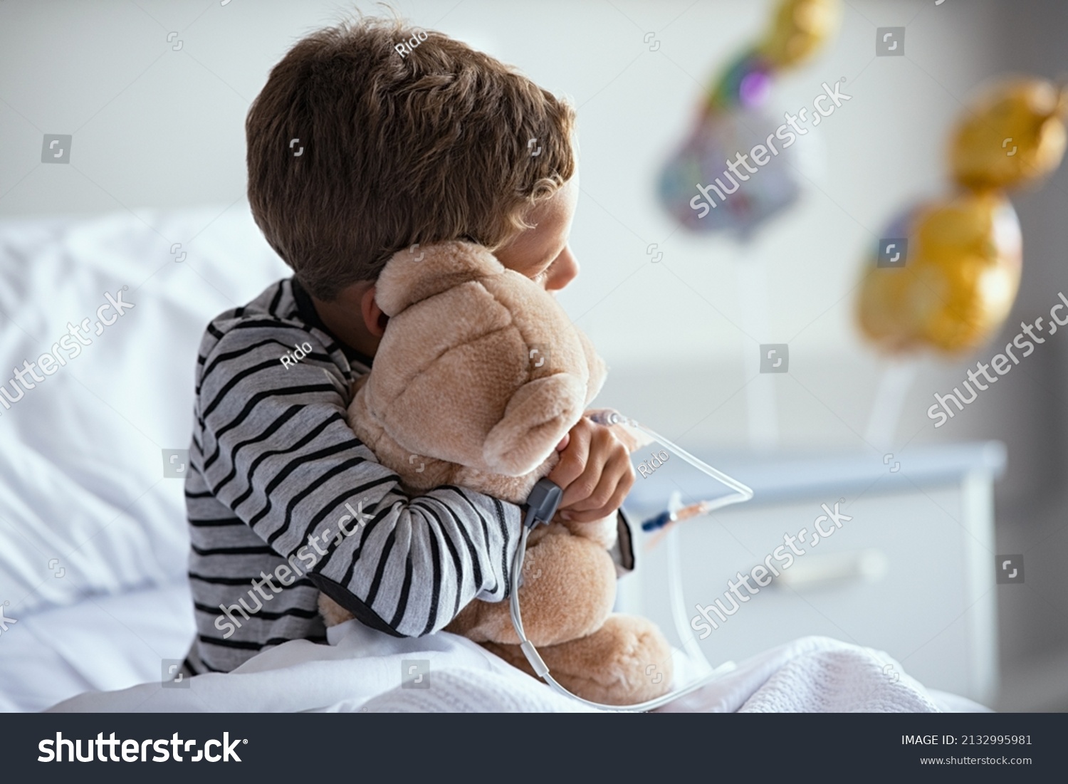 Rear view of scared little boy with intravenous drip in hand hugging teddy bear sitting on hospital bed. Sick lonely child looking through the window in clinic pediatric ward before the surgery. #2132995981