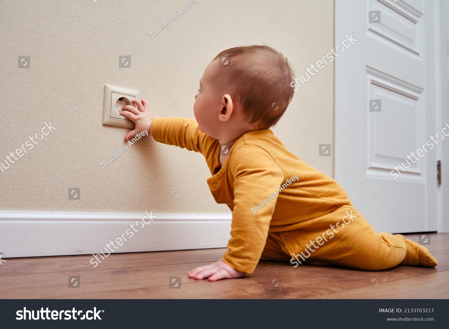 Baby toddler reaches into the electrical outlet on the home wall with his hand. Danger and protection of child fingers from electric shock, aged 6-11 months #2133703217