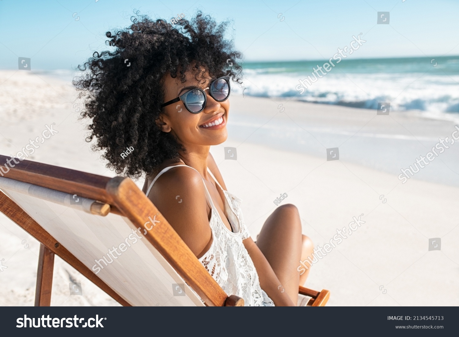 Portrait of happy young black woman relaxing on wooden deck chair at tropical beach while looking at camera wearing spectacles. Smiling african american girl with fashion sunglasses enjoying vacation. #2134545713