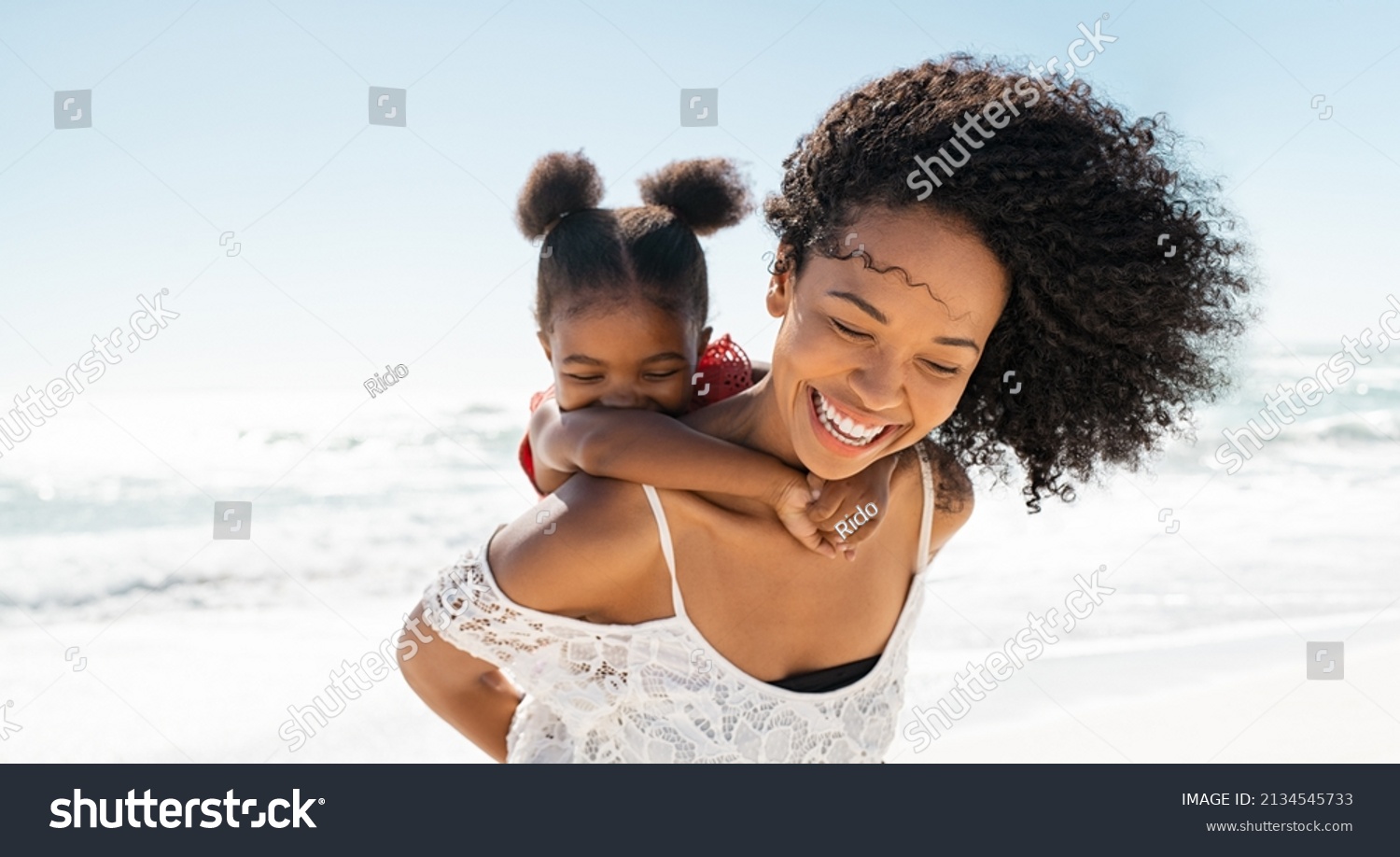 Happy black mother giving piggyback ride to her daughter at beach while laughing. Close up of beautiful and carefree mom carrying on shoulders her kid. Cute african little girl with bigger sister play #2134545733