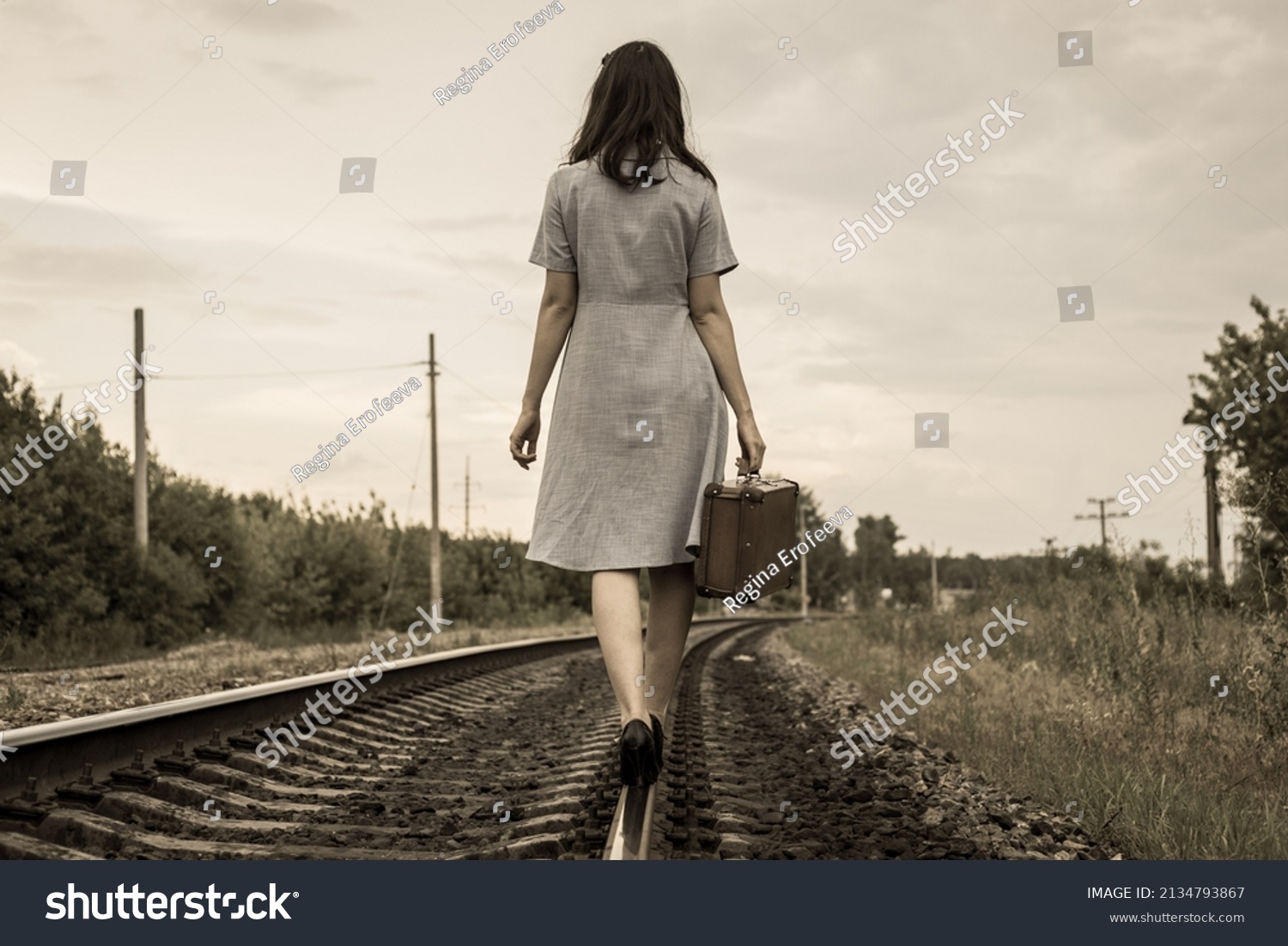 A rear view of a young woman in a dress and with a suitcase walking away along the rails of a railway road. The concept of departure, emigration, departure from the country, refugee status,deportation #2134793867