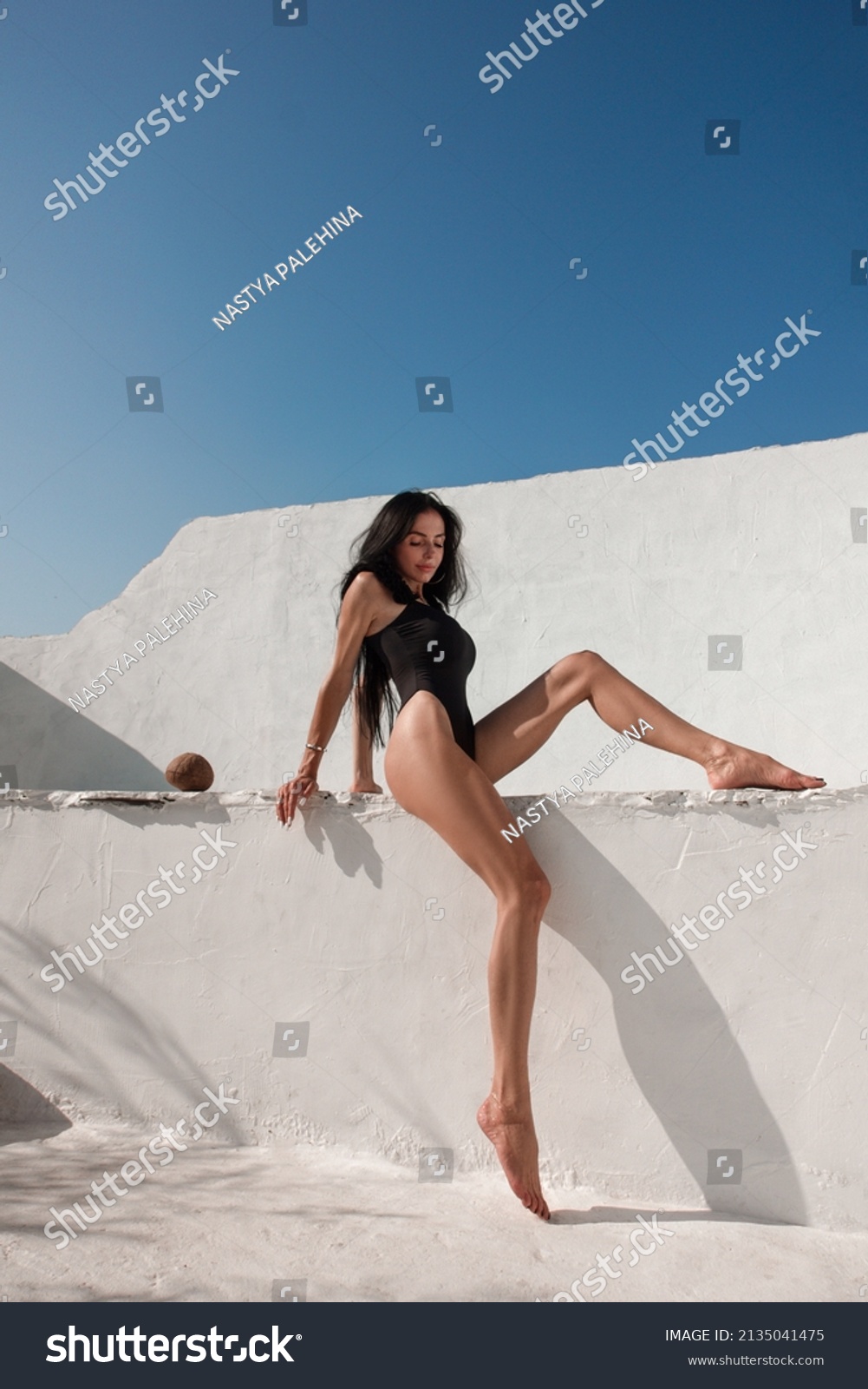 vacation mood. brunette woman with long legs in black glamour swimsuit sits fashion on the  swimming pool with coconut near white wall background at sunny day. greek villa. travel concept, free space #2135041475