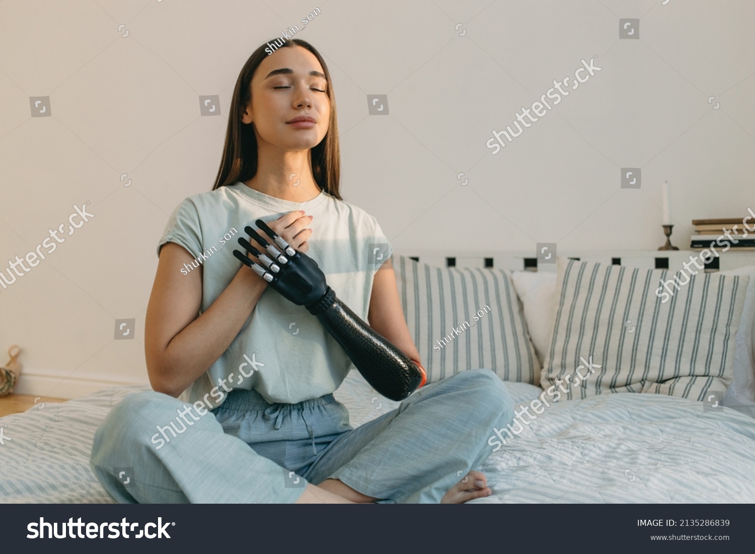 Young cyborg female practicing yoga in bed in early morning, meditating, keeping hands on chest, breathing, having bionic black prosthesis instead of left hand, wearing cute cotton pajama #2135286839