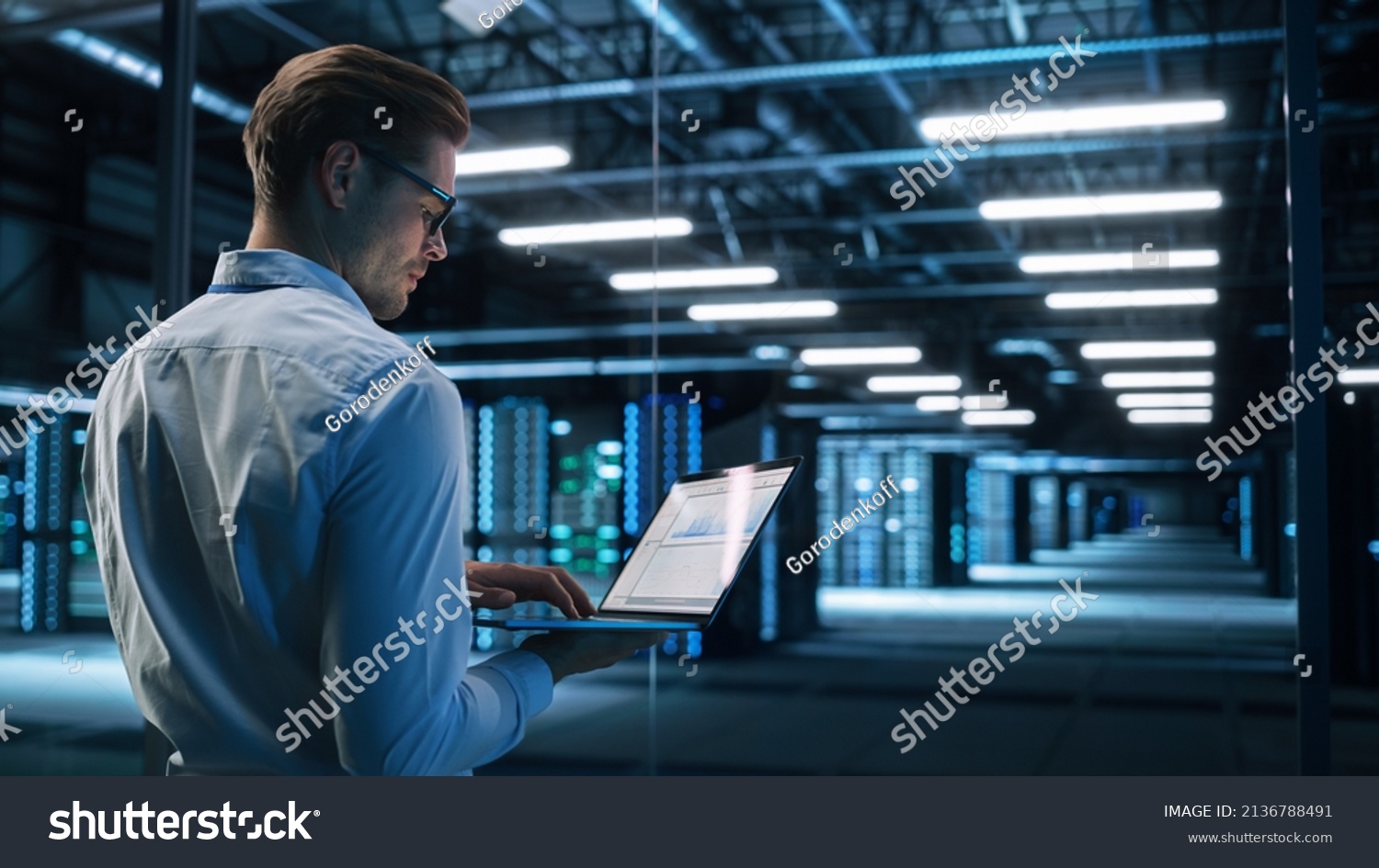 Back View of Male Specialist Using Laptop in Big Data Center Office. High Speed Data Transfer, Server Transfer, Technology Science Breakethrough, Progress, Innovation Concept #2136788491
