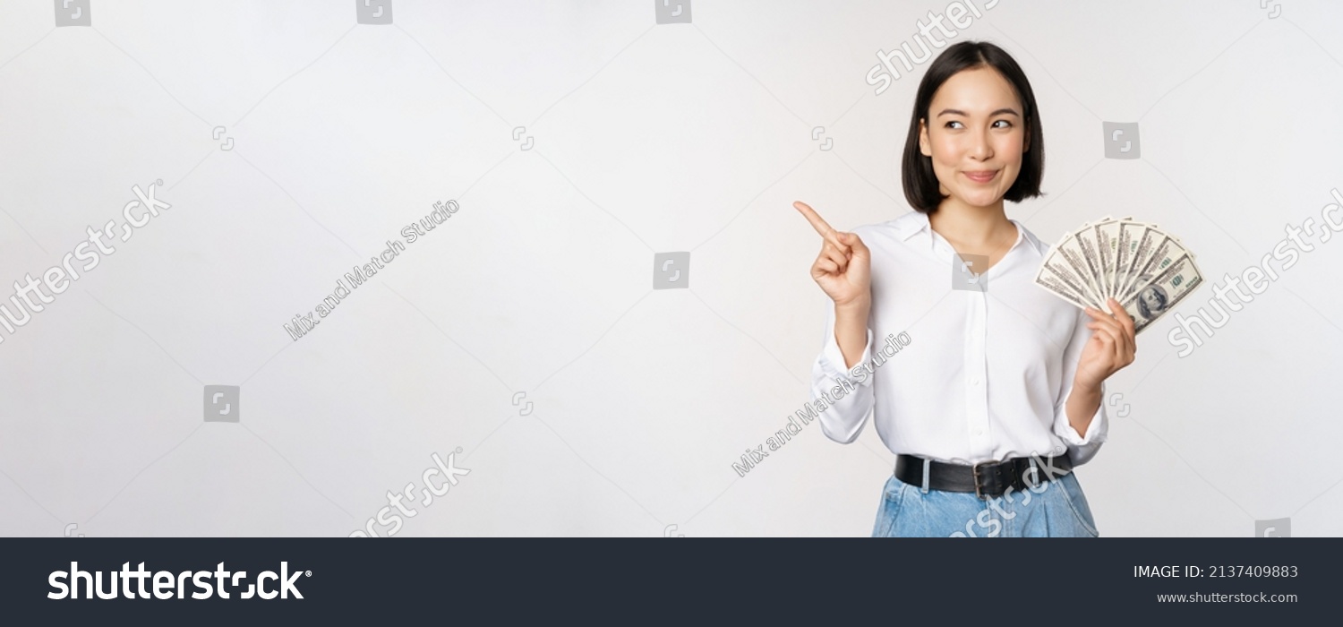 Smiling young modern asian woman, pointing at banner advertisement, holding cash money dollars, standing over white background #2137409883