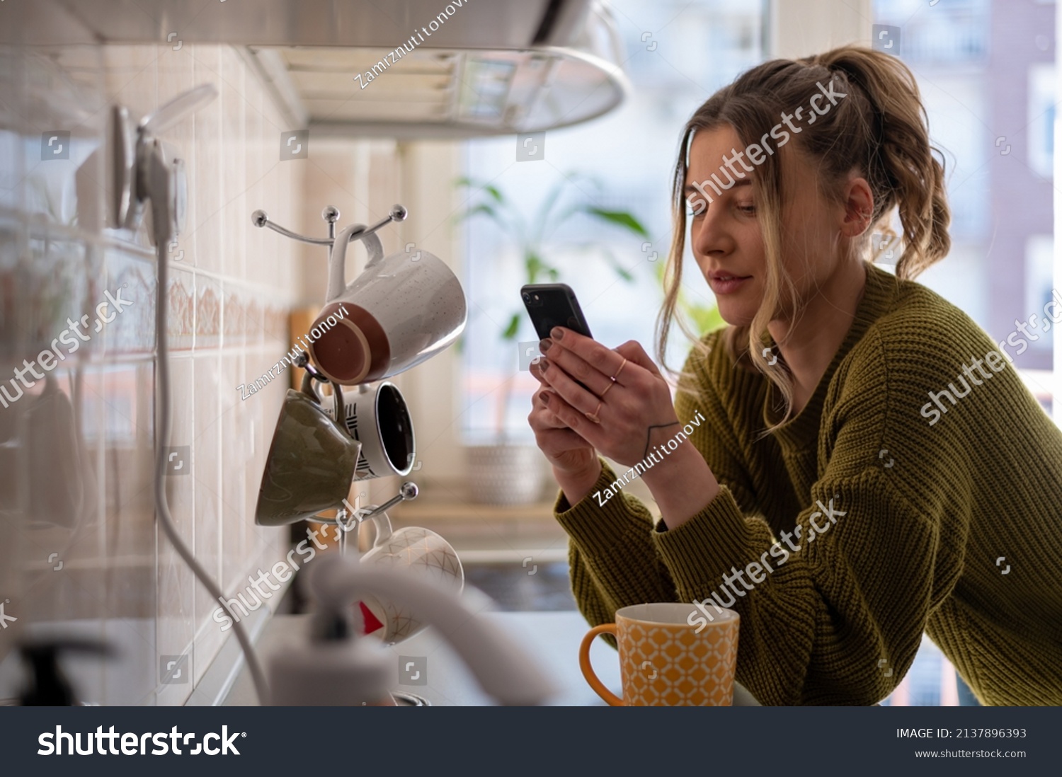 Shot of a young woman drinking tea and using smartphone in her kitchen while getting ready to go to work. Watching videos or messaging with friends while enjoying morning at home. #2137896393