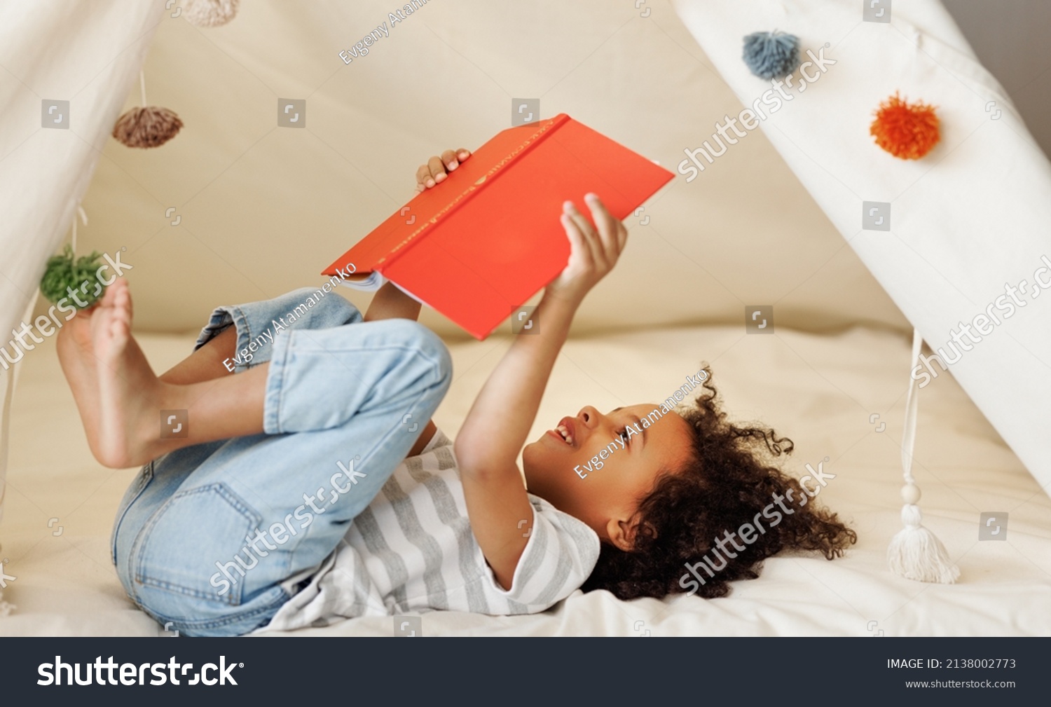 Portrait of cute little african american kid curly boy with book smiling at camera while   reading book  in play tent at home, happy child playing alone in children room #2138002773