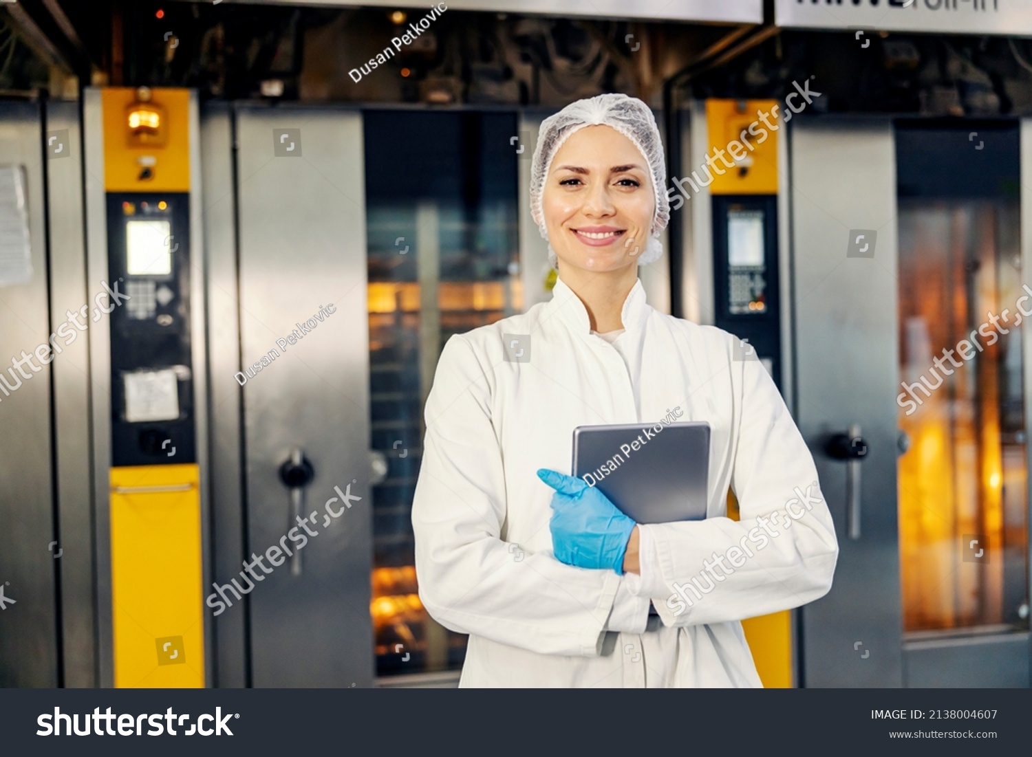 A smiling female food factory inspector with tablet in hands standing in front of ovens in bakery. #2138004607