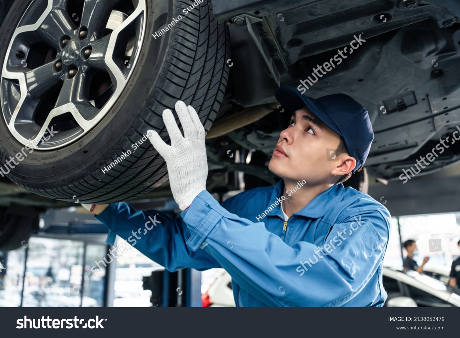 Asian automotive mechanic repairman look under car condition in garage. Vehicle service guy worker doing check or mend car wheel and work in mechanics workshop with confidence to repair car engine car #2138052479