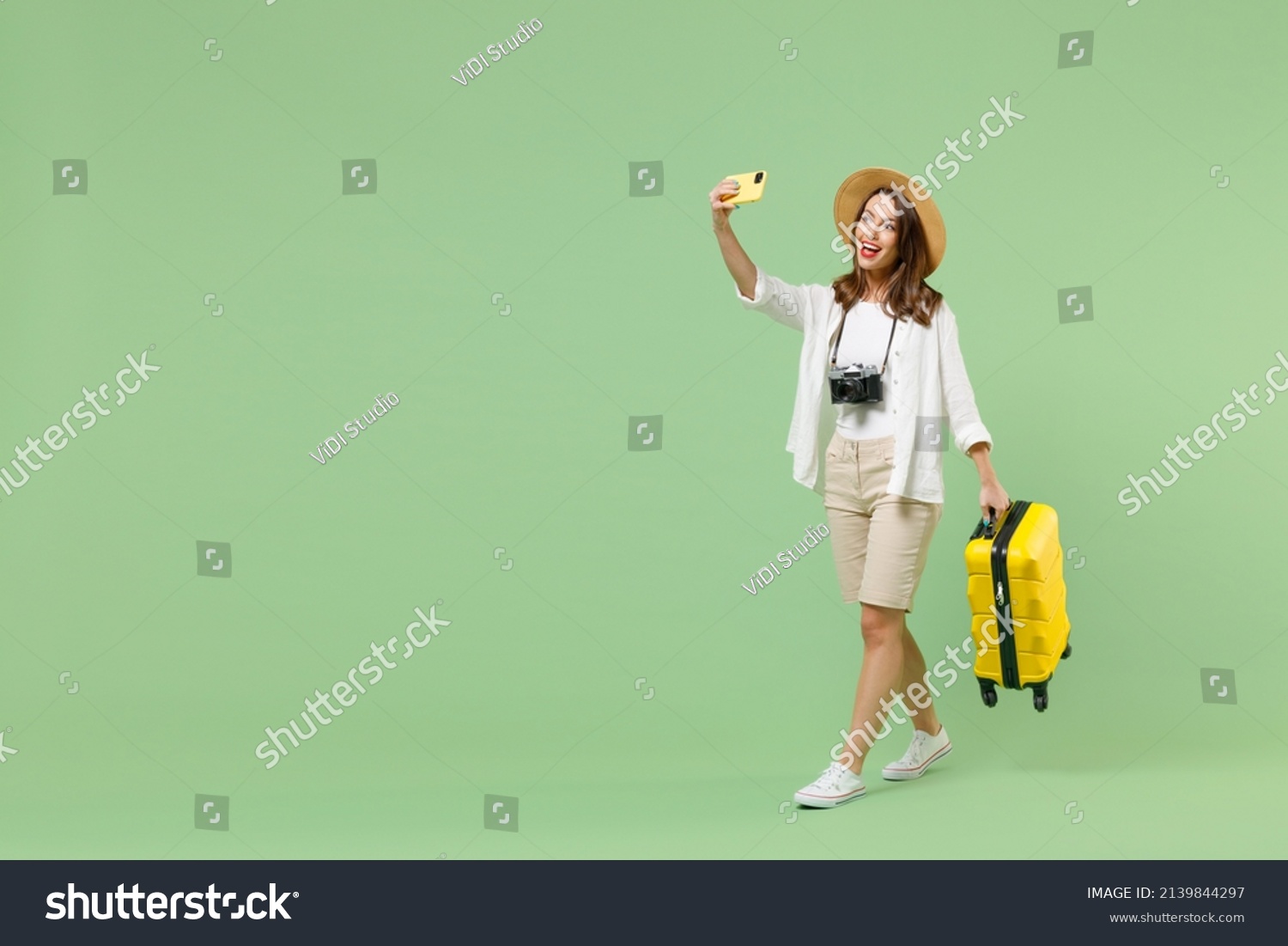 Full length traveler tourist woman in casual clothes hat hold suitcase do selfie on mobile cell phone isolated on pastel green background. Passenger travel abroad weekends. Air flight journey concept #2139844297