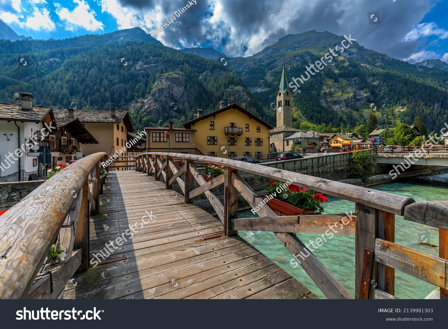 Wooden bridge over mountain river as houses and old church on background in small town of Gressoney-Saint-Jean in Aosta Valley, Italy. #2139981303