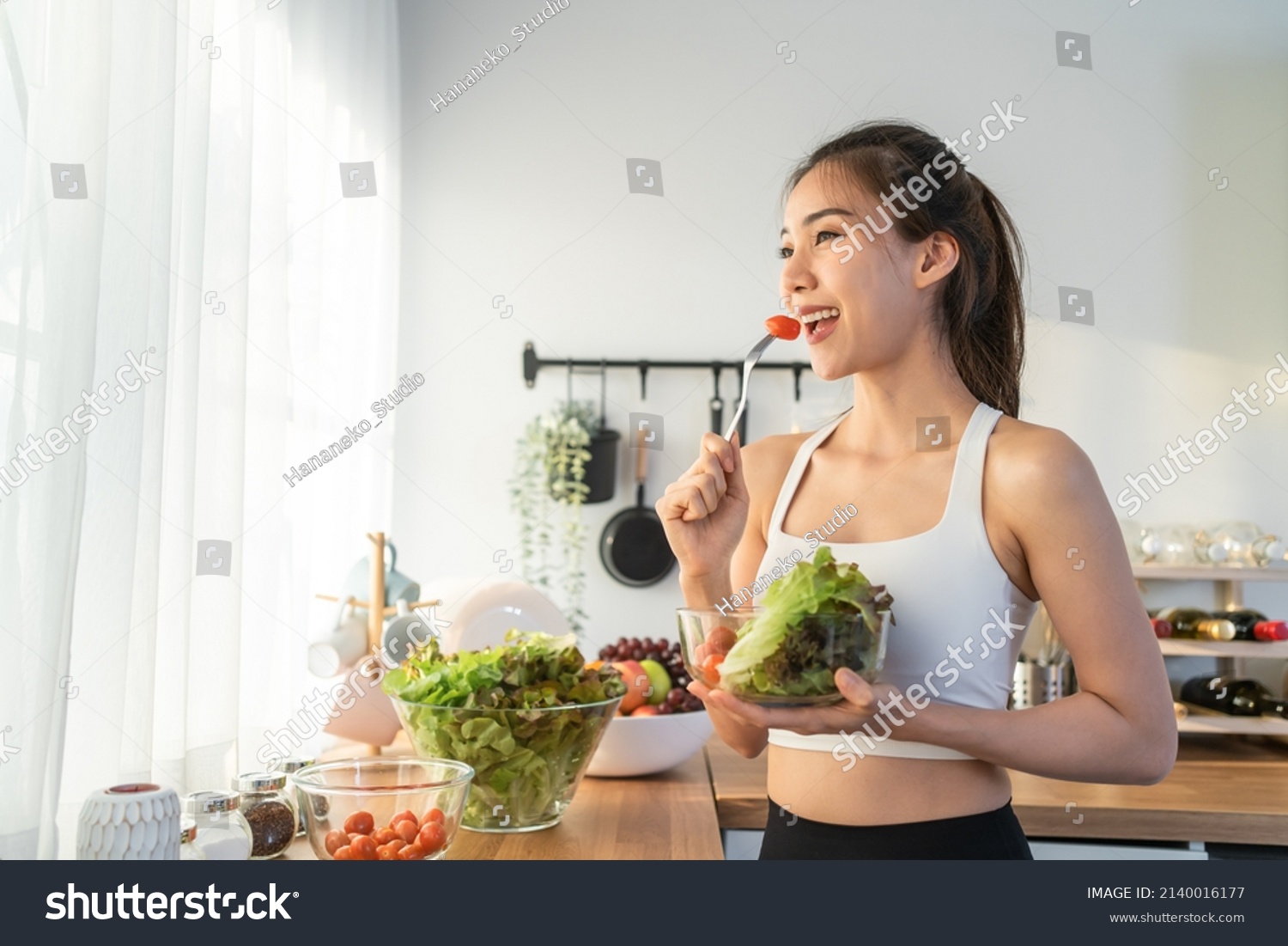 Asian attractive sport woman holding salad bowl and eat vegetables. Beautiful sport girl in sportswear enjoy eat clean vegetables after exercise for health in house. Diet and Healthy food concept. #2140016177