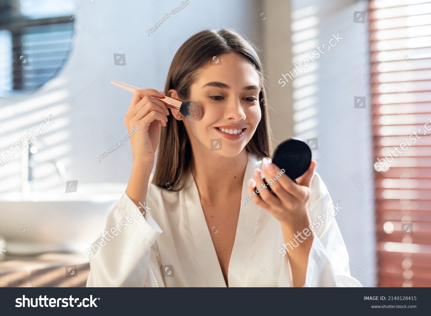 Daily Makeup. Smiling Young Female Applying Blush With Makeup Brush In Bathroom, Beautiful Happy Woman Wearing White Silk Robe Using New Cosmetics While Getting Ready At Home, Copy Space #2140128415