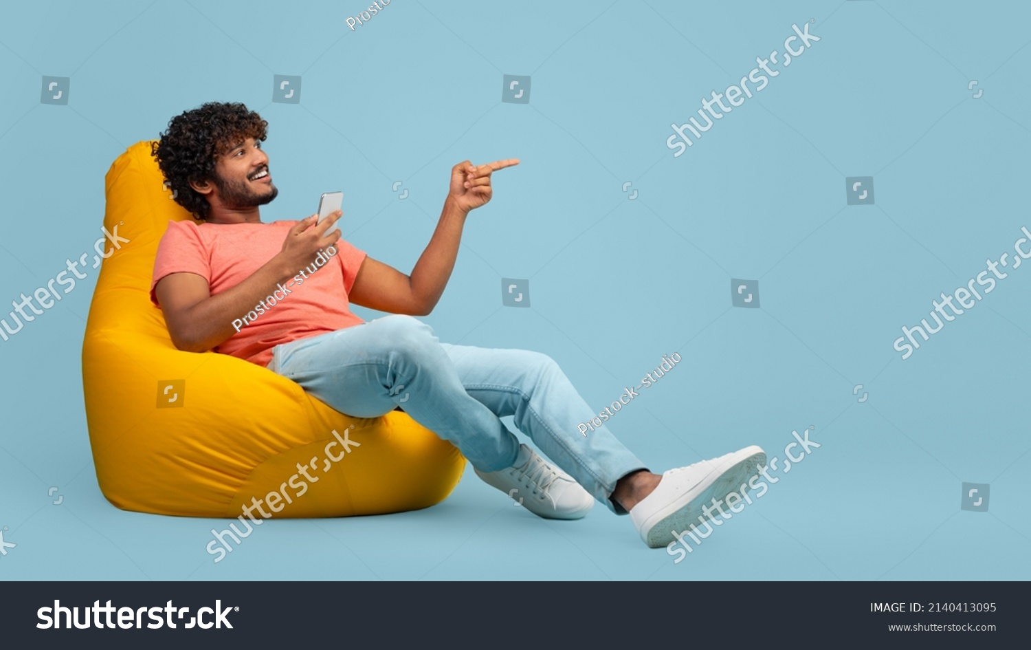 Happy curly millennial indian man sitting at bean bag with brand new smartphone and pointing at copy space over blue studio background, showing nice offer and smiling, panorama #2140413095