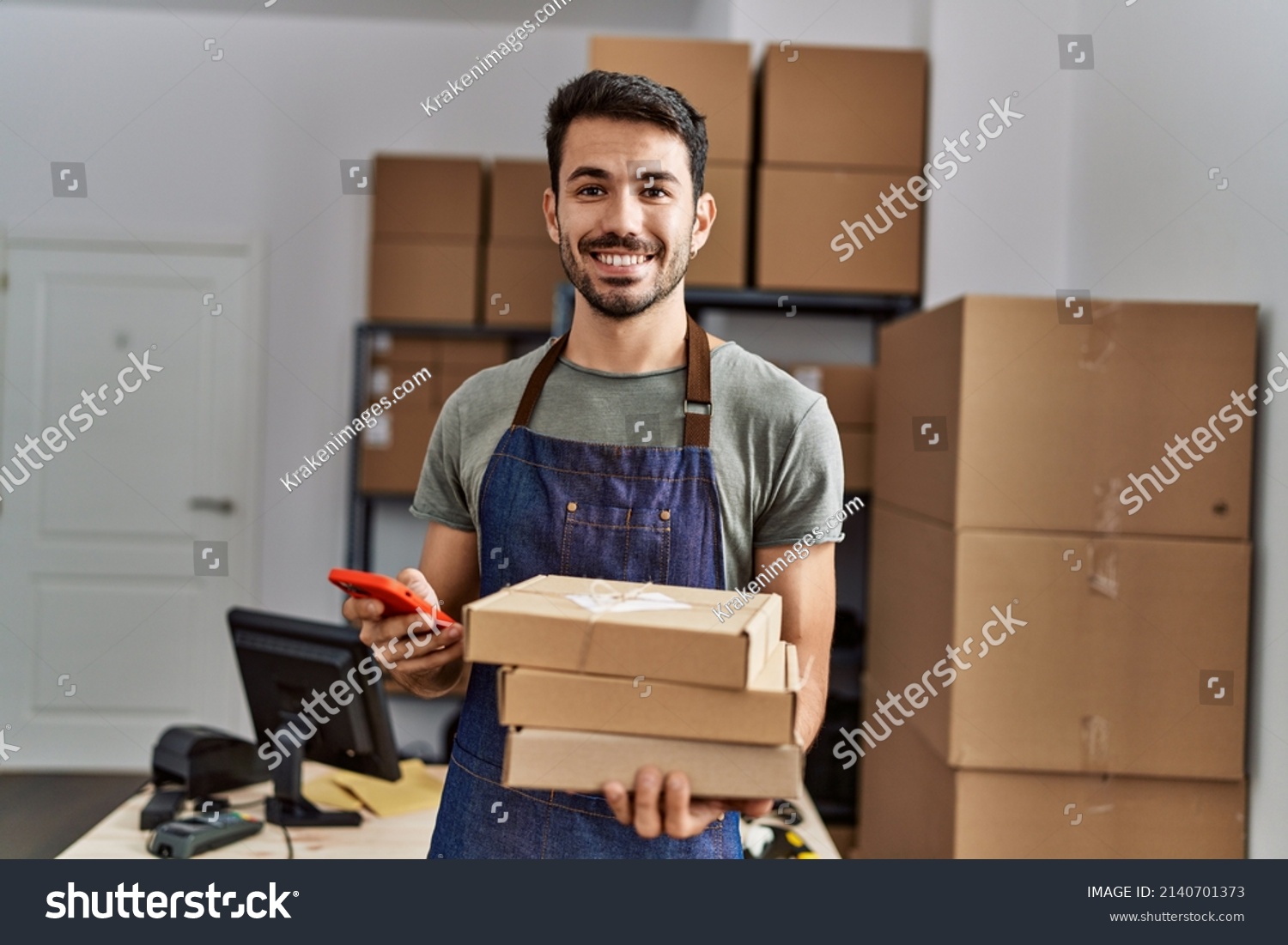 Young hispanic man business worker using smartphone holding packages at storehouse #2140701373