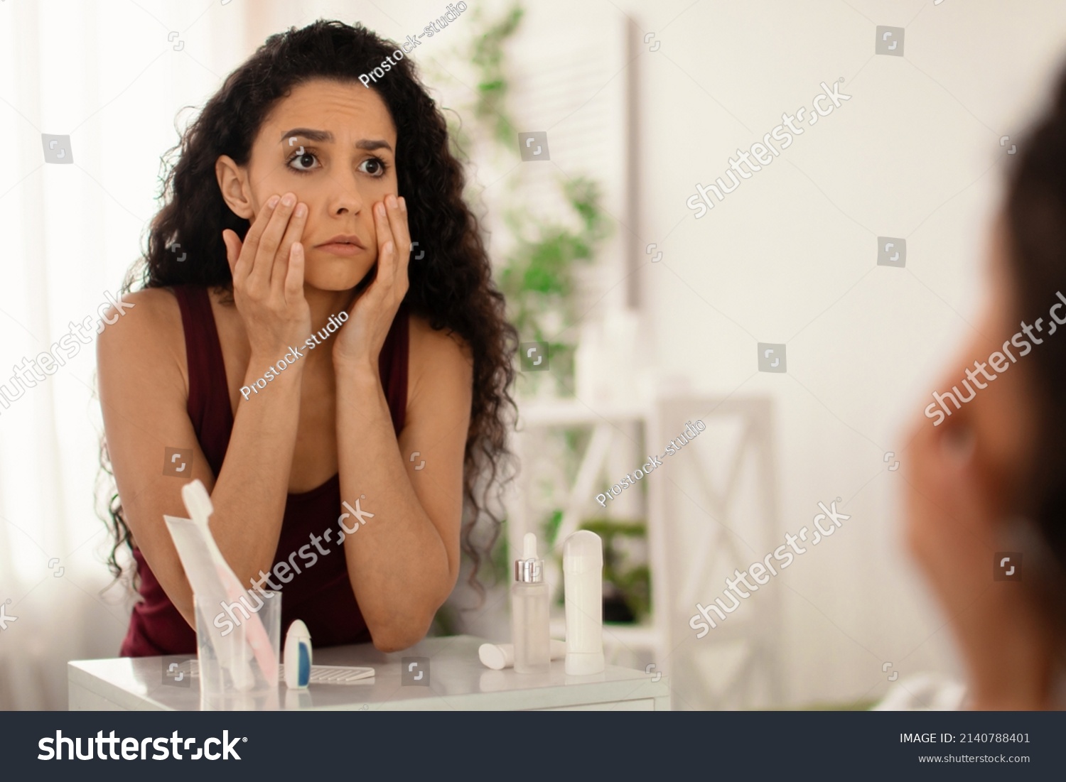 Beauty and skincare. Upset young woman looking in mirror and touching face, examining wrinkles and dark circles under her eyes at home, copy space. Dull tired skin concept #2140788401