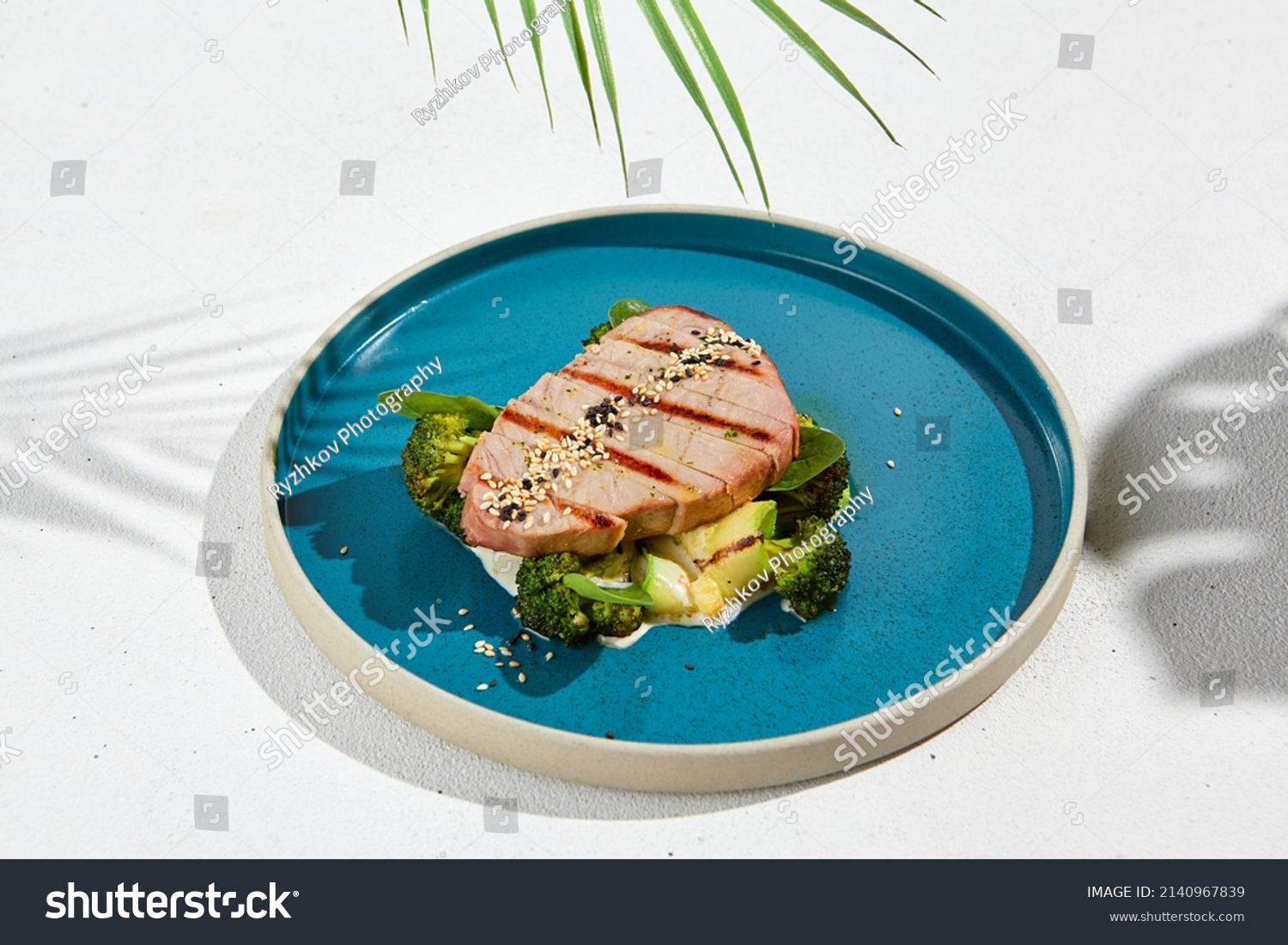 Grilled tuna steak with broccoli and zucchini in modern ceramic plate. Healthy food - roasted tuna with green vegetables. Fish dish in minimal style. Tuna fillet in blue plate with hard shadow #2140967839