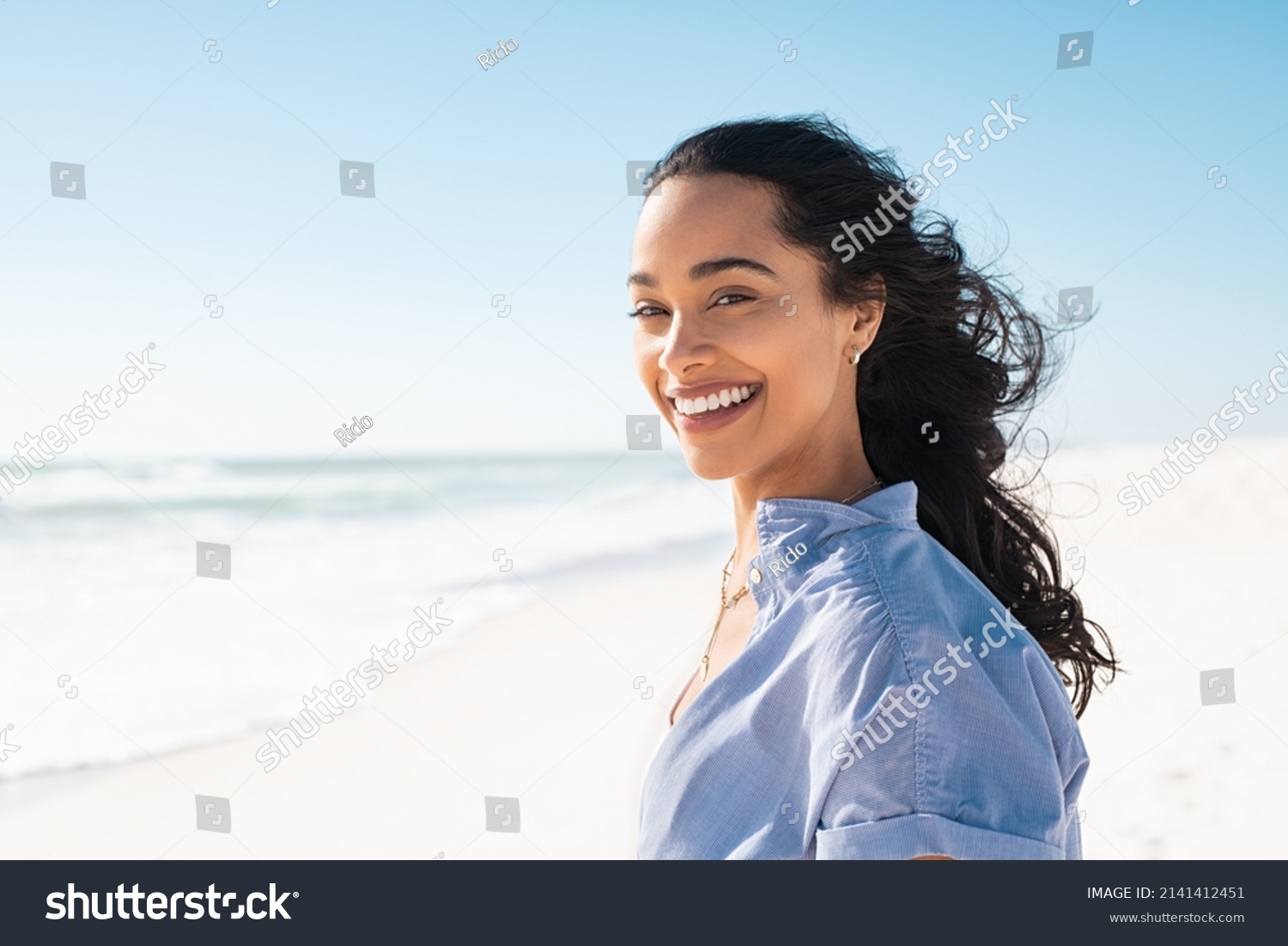 Portrait of young woman at sea looking at camera. Smiling latin hispanic girl standing at the beach with copy space and looking at camera. Happy mixed race girl in casual outfit with wind in her hair. #2141412451