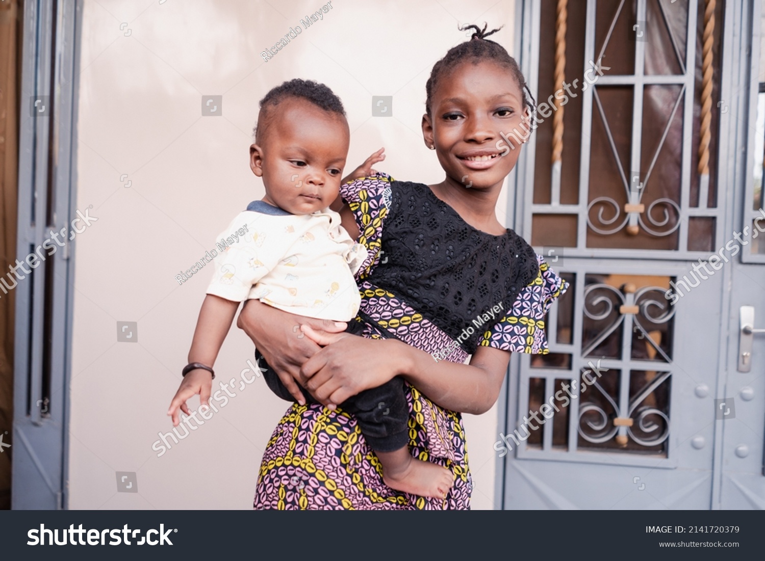 Responsible proud big sister takes care of her little brother, whom she carries in her arms as a matter of course according to African tradition and family ties #2141720379