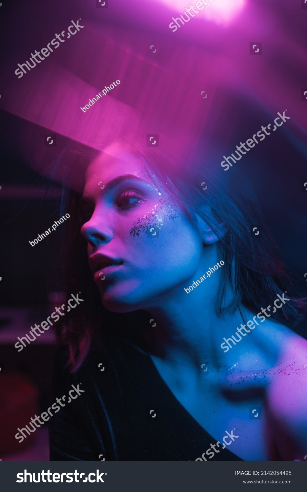 Fashionable photo of a woman with bright makeup in neon light with purple and blue in a dark room, looking away. vertical #2142054495
