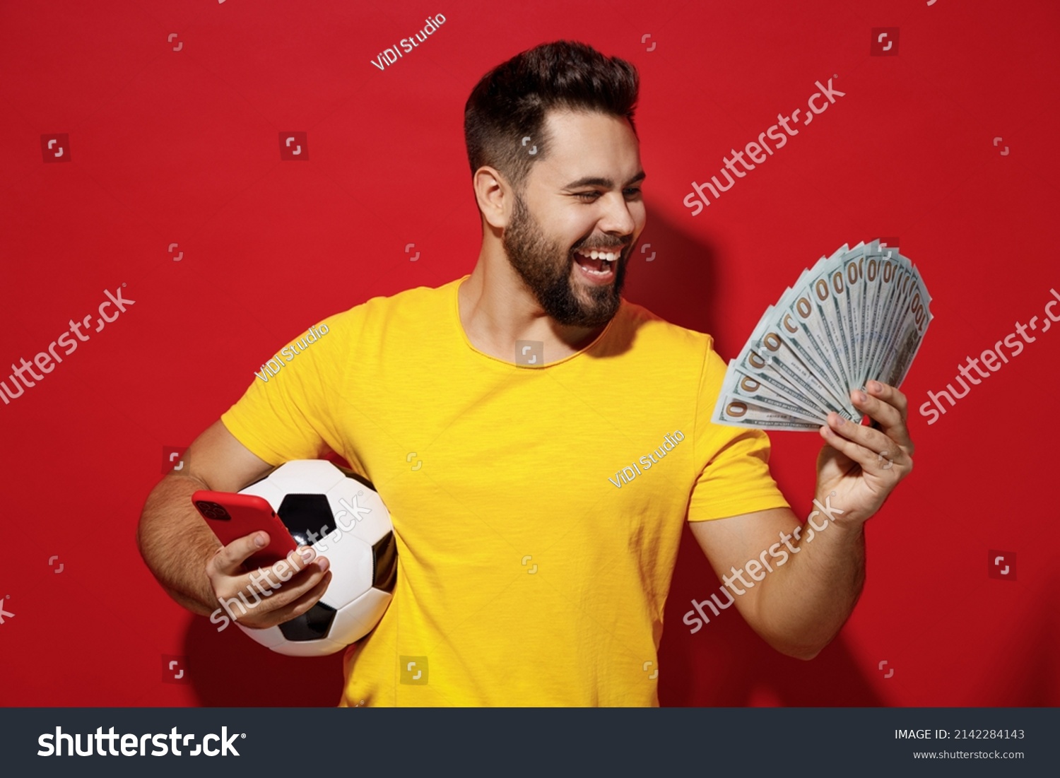 Fun young bearded man football fan in yellow t-shirt cheer up support favorite team hold soccer ball look at fan of cash money in dollar banknotes isolated on plain dark red background studio portrait #2142284143