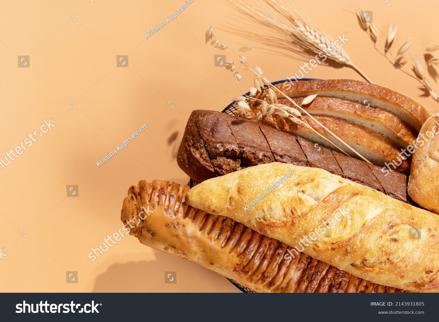 Homemade natural breads. Fresh loafs of bread in the blue basket with ears of rye and wheat on a beige background with copy space. Crunchy french baguettes, slices of bread and a bun. Soft focus  #2143931805