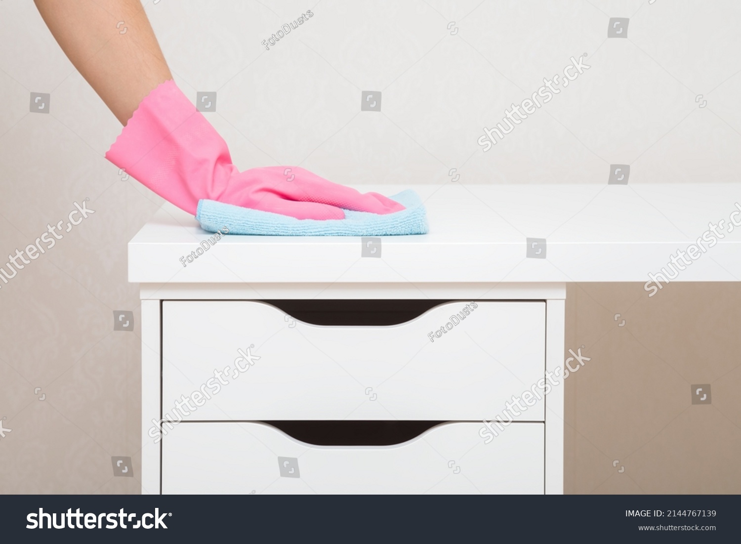 Young woman hand in pink rubber protective glove holding blue dry rag and wiping dust from white work table with drawers at home office. Closeup. General or regular cleanup in room. Cleaning service. #2144767139