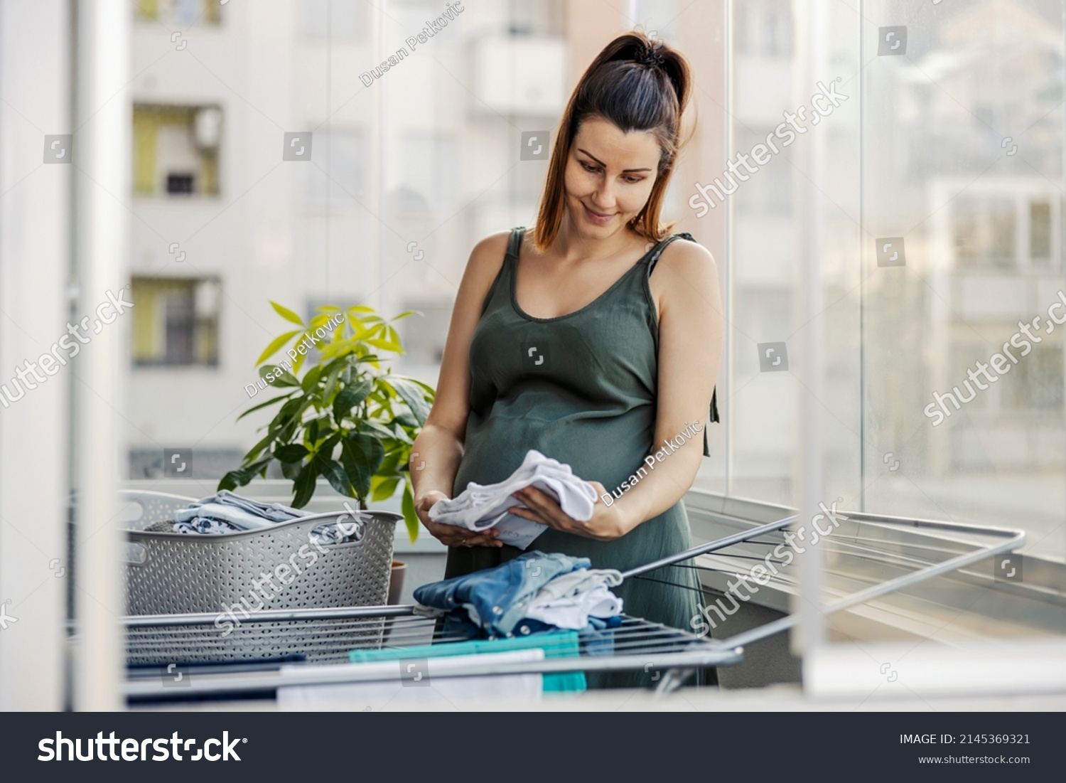 Maternal instinct, house chores and pregnant women. An excited expectant mother prepares clothes for the newborn. A pregnant woman stands on the terrace and prepares clothes for the newborn #2145369321