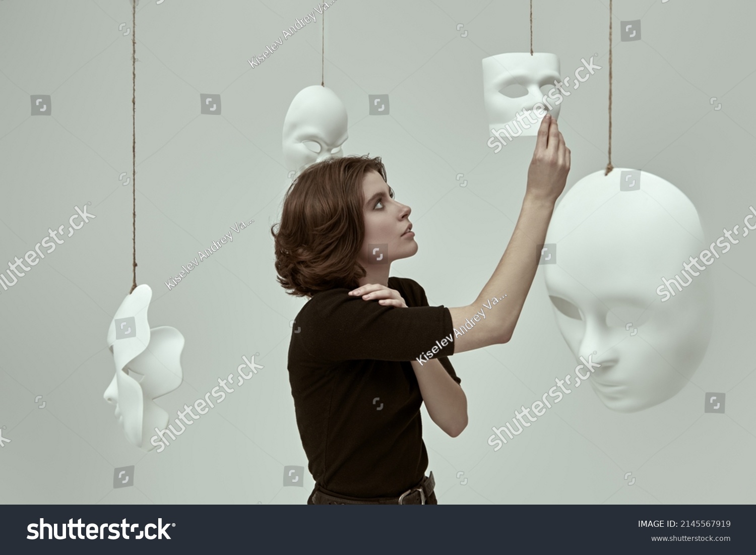 Human roles. A girl in black clothes stands in a white room among different masks deciding which one to choose. Hypocrisy. Mental disorders. #2145567919