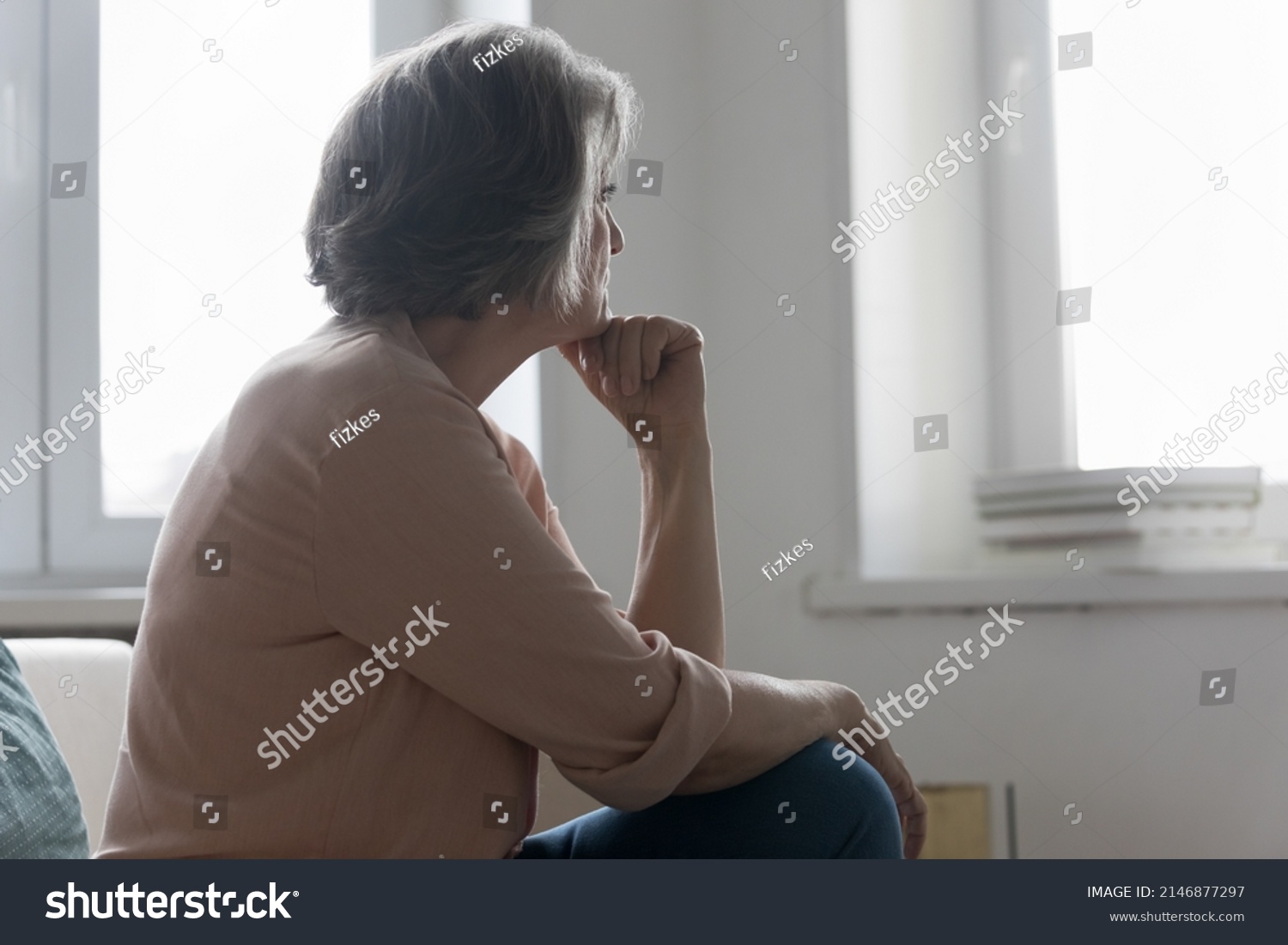 Serious older 50s woman looks out window spend time alone at home, feels sad or disappointed, waits or misses someone seated on sofa indoor. Loneliness, solitude on retirement, nostalgic mood concept #2146877297