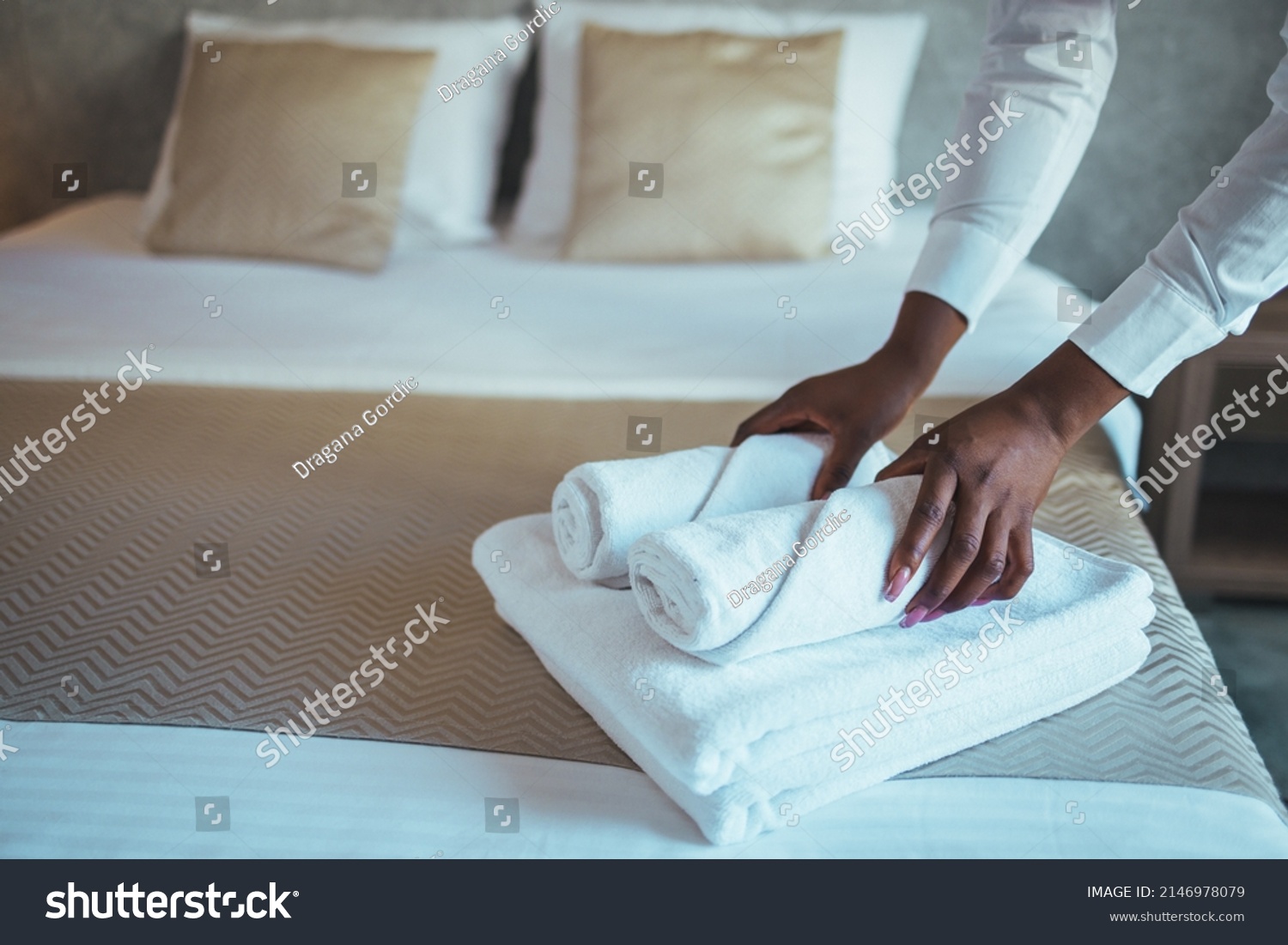 Young beautiful chambermaid in uniform holding stack of clean folded towels while going to put them on changed bed in hotel room.  Photos in the interior. #2146978079