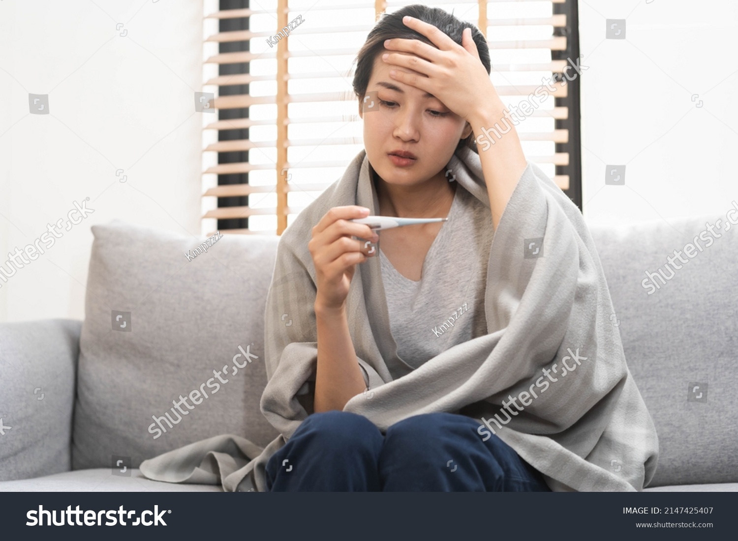 Sick, influenza asian young woman, girl headache have a fever, flu and check thermometer measure body temperature, feel illness sitting on sofa bed at home. Health care person on virus, covid-19. #2147425407