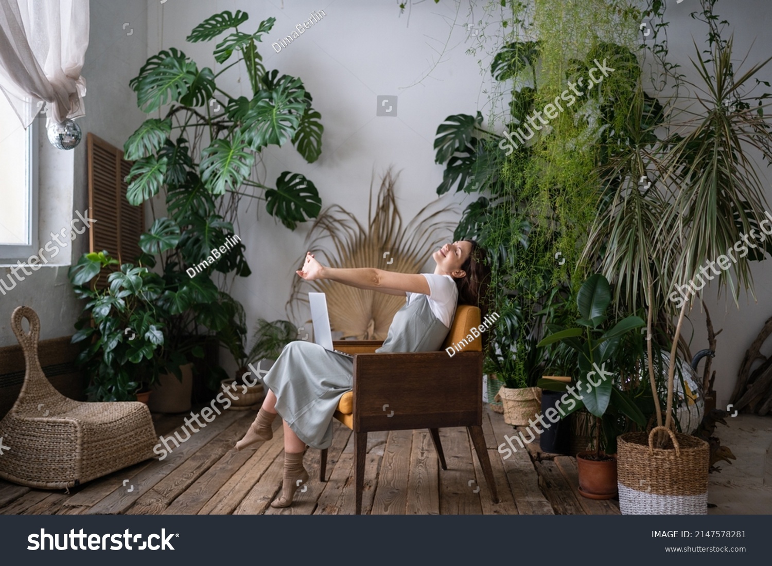 Overworked woman freelancer sitting on chair in cozy greenhouse, resting, stretching arms with closed eyes. Smiling tired woman in home garden taking break from online study. Plant lovers concept. #2147578281