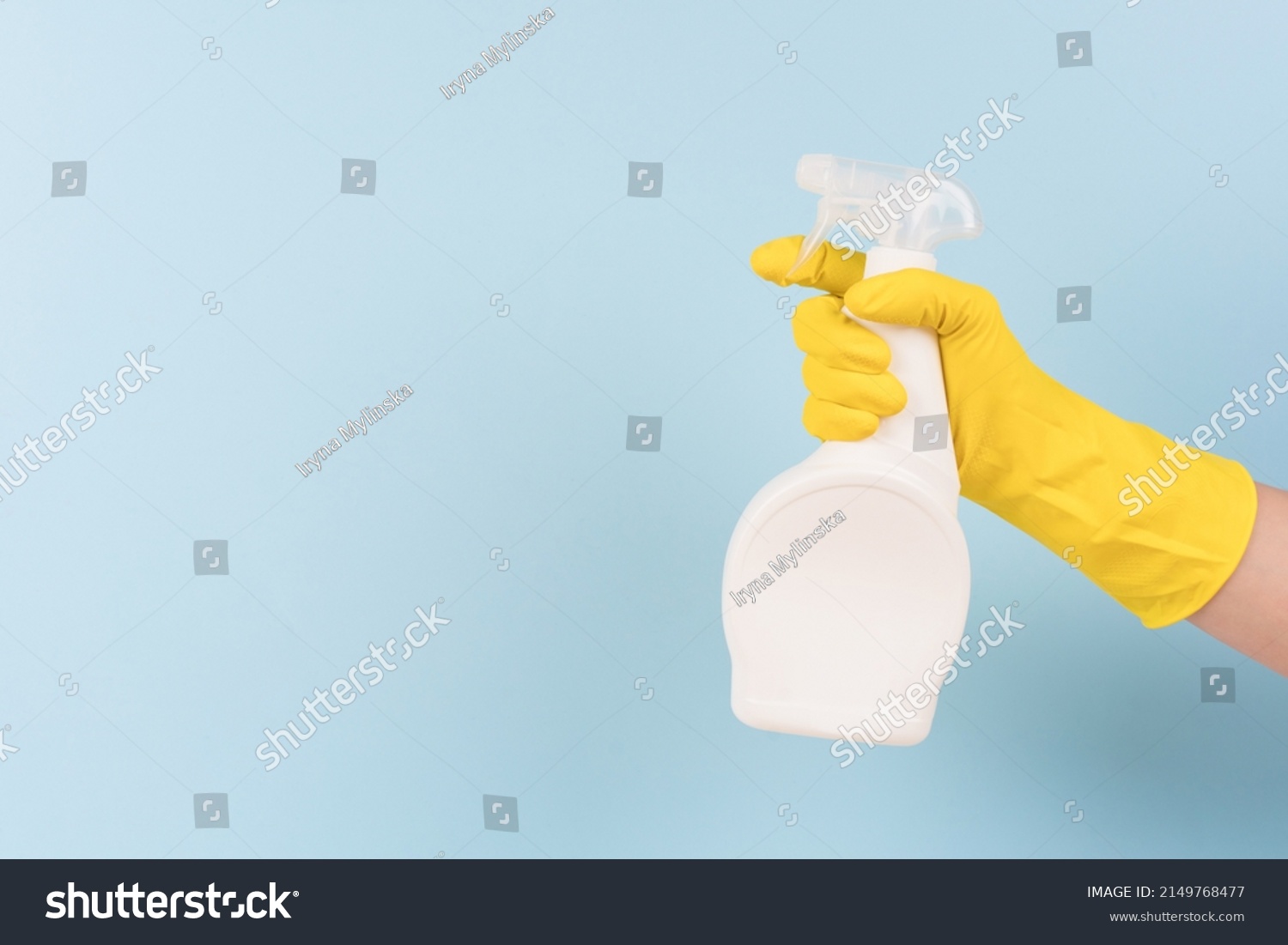 Hand in yellow protective rubber glove holding spray bottle with detergent against blue background with copy space. Cleaning product for different kitchen or bathroom surfaces. Bleach. Mockup #2149768477