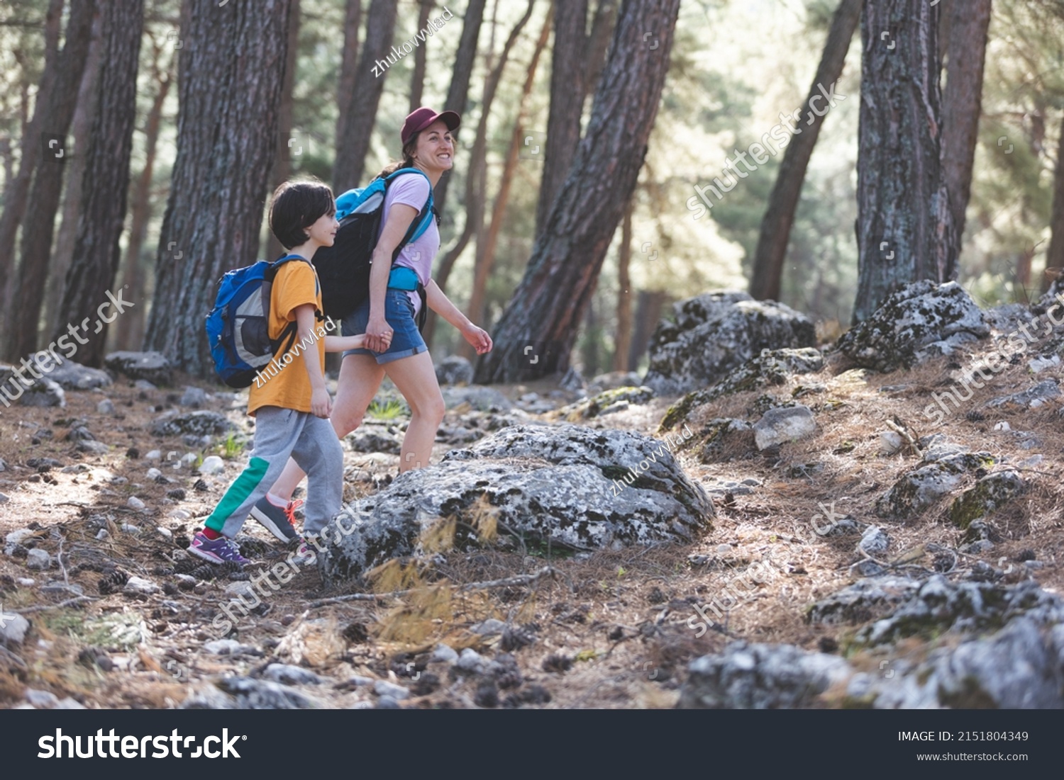 A woman walks with her son through the forest, The boy with his mother go hiking, A child with a backpack is in the park, Travel with children, The kid holds mom's hand, Mountain trail, Mothers Day. #2151804349