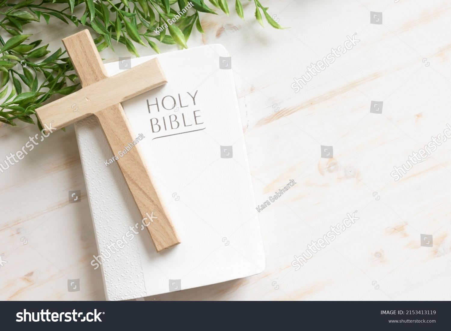 Wood cross laying on a closed white Christian bible with a green vine border all on a white wood background with copy space #2153413119